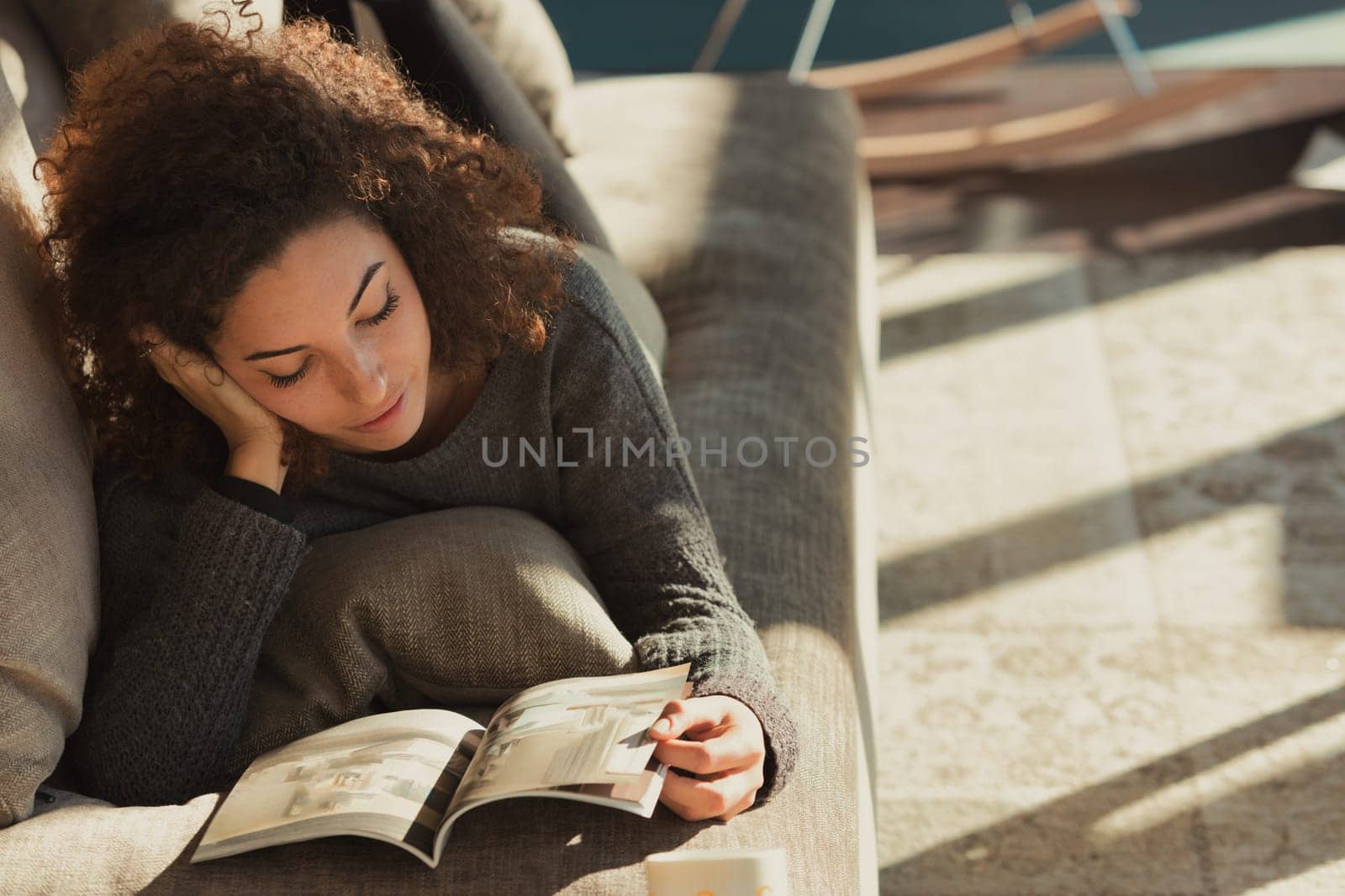 A relaxed young woman with curly hair lies belly-down on the couch in her spacious, sunlit living room, reading a magazine or catalog while enjoying her free time and having fun. Home sweet home