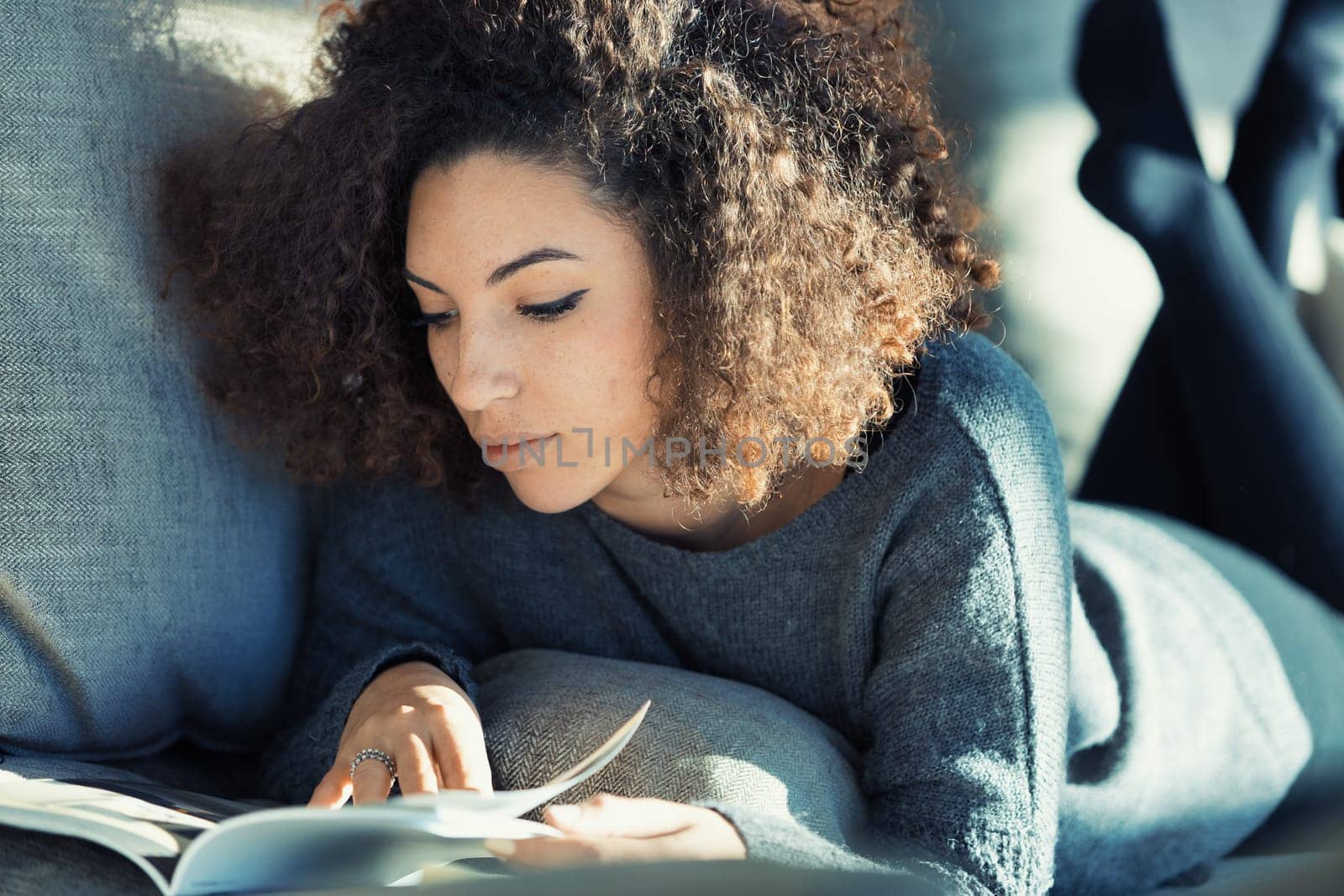 A stunning curly-haired model lounges on her sofa, immersed in reading a magazine or catalog. With her legs crossed and up behind her, she's barefoot. The sun kisses her Afro hair and her dress harmon
