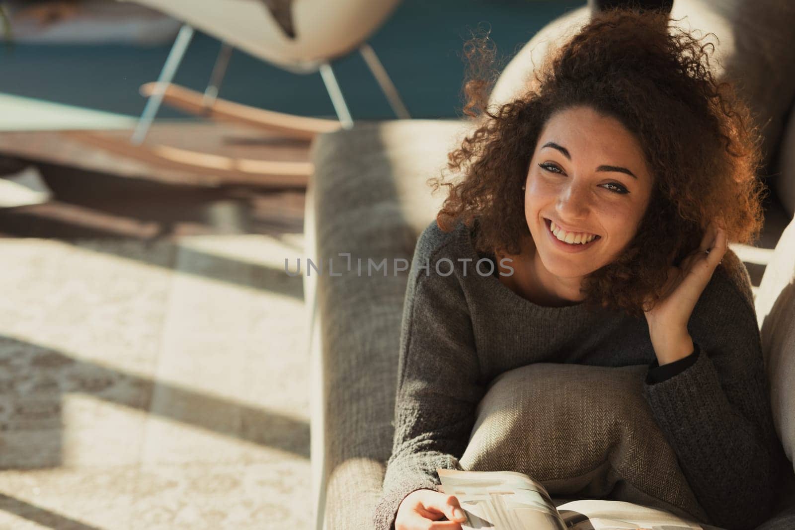 A relaxed young woman with curly hair is sprawled out on her stomach on the couch in her spacious and bright living room bathed in sunlight. She is reading a magazine or catalog with her head resting
