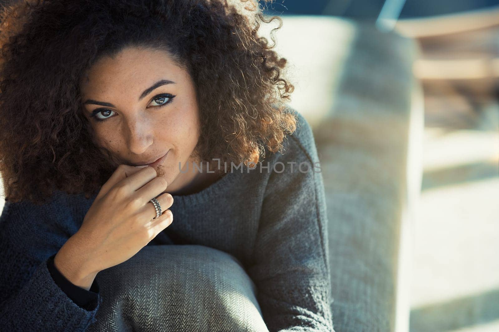 In her sunlit living room, a curly-haired young model lounges on her sofa, reading with one hand at her lips. Her captivating eyes and adorable coy smile radiate charm, as we admire her perfectly mani