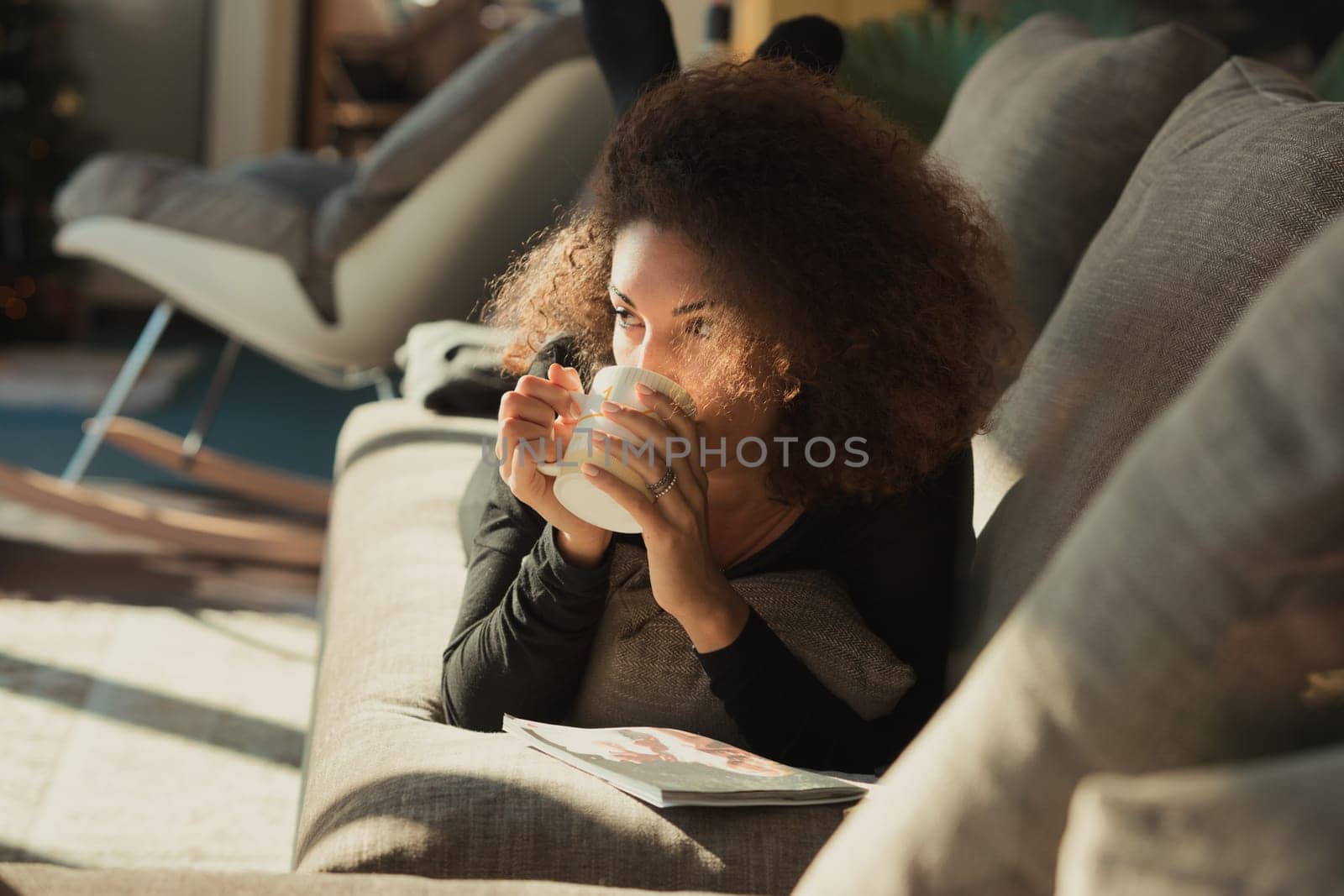 beautiful curly-haired woman is engrossed in reading a magazine. She takes a moment to thoughtfully sip a hot beverage, perhaps a herbal tea or regular tea, from her mug. She is comfortably settled on