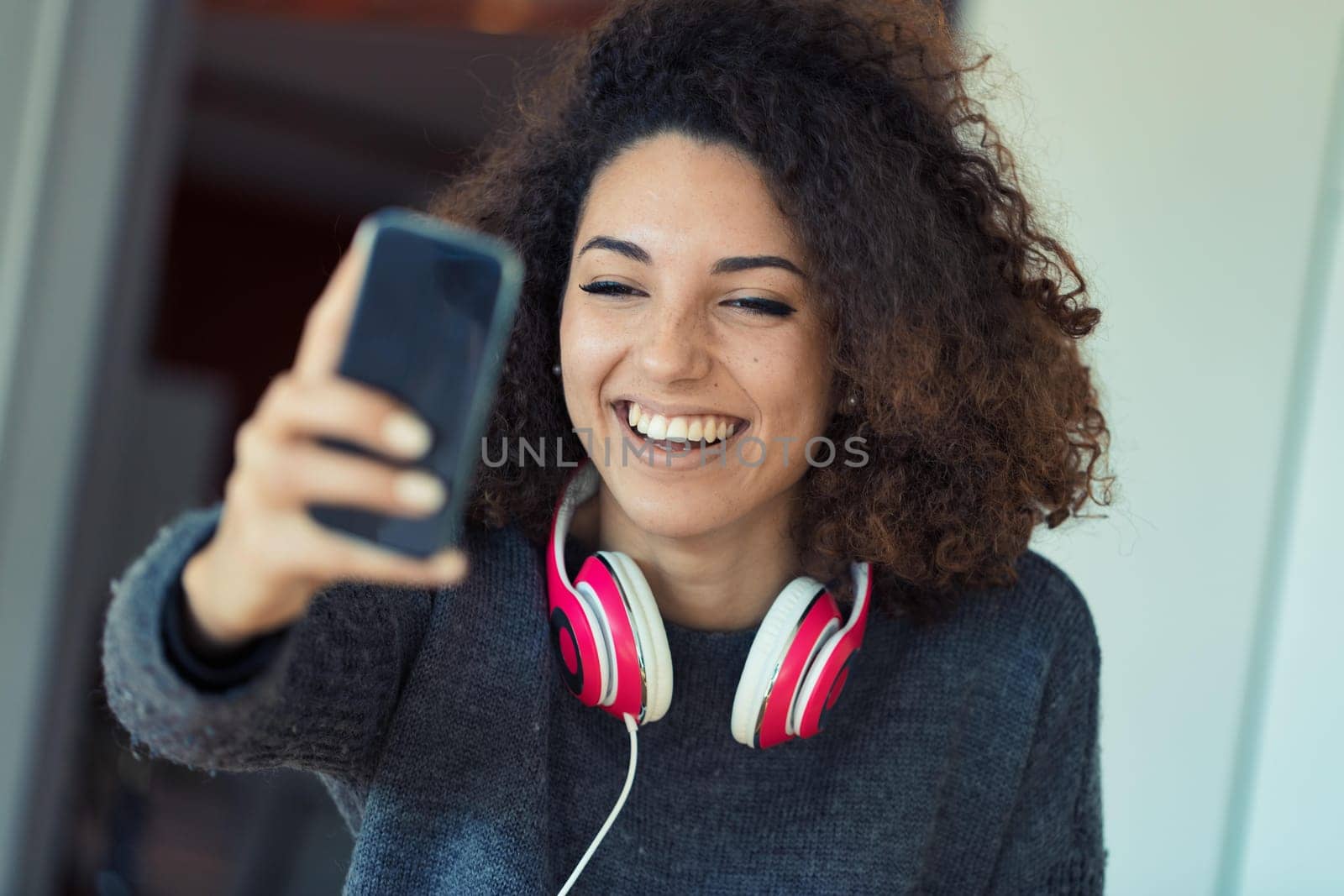 A young, freckled, curly-haired woman in a video call. Laughing and enjoying, she wears pink headphones for privacy. Happy student or worker with perfect teeth