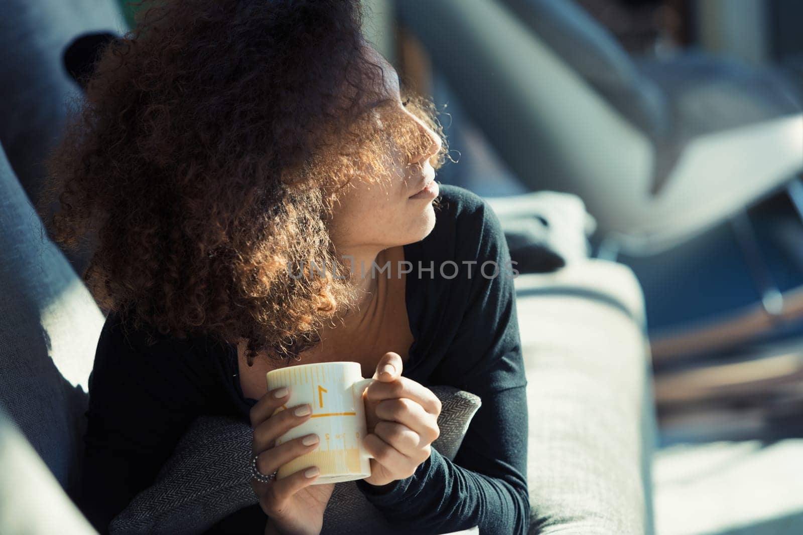 An affluent young Afro-American woman with curly hair lies belly-down on her own couch, savoring a cup of tea. She's facing the sunlight that beams through the window of her sizeable, stylish living r