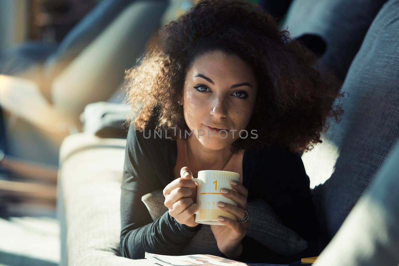 Relaxing in her living room, a curly-haired model lounges with a warm drink. Sunlight bathes her as she basks in its glow. Her beautifully manicured hands hold our attention as her enchanting eyes mee
