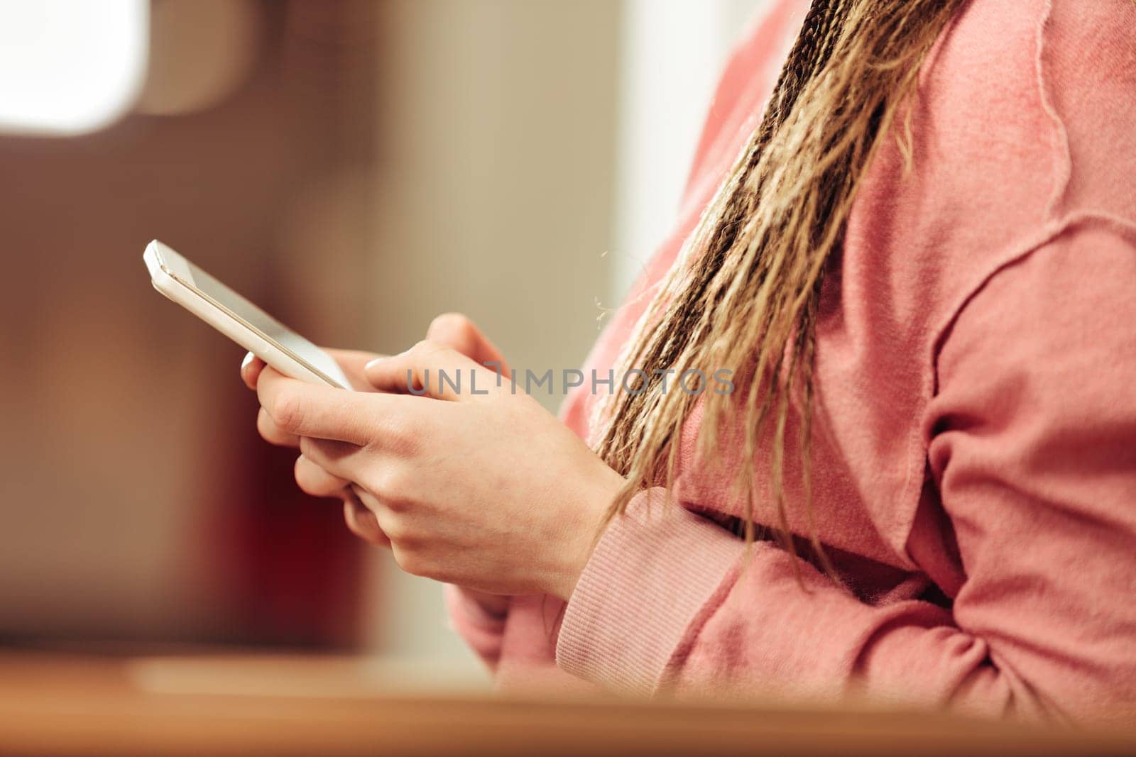 In warm light, close-up detail of female hands with smartphone at home with blurred background. She has an orange sweatshirt and box-braids