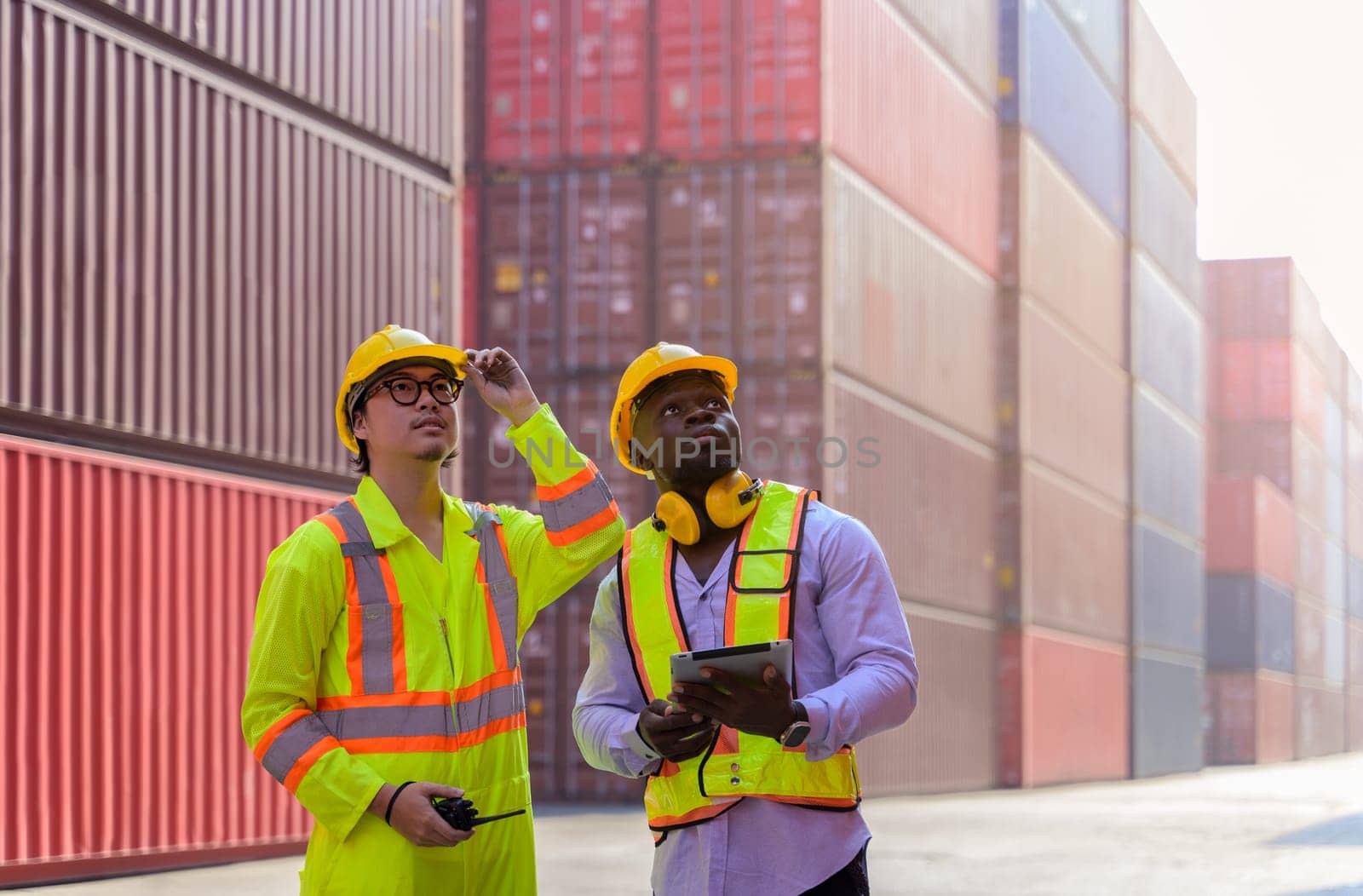 Two men standing at cargo shipping yard and checking on loading containers . Asian man worker holding walkie-talkie while African engineer holding digital tablet.