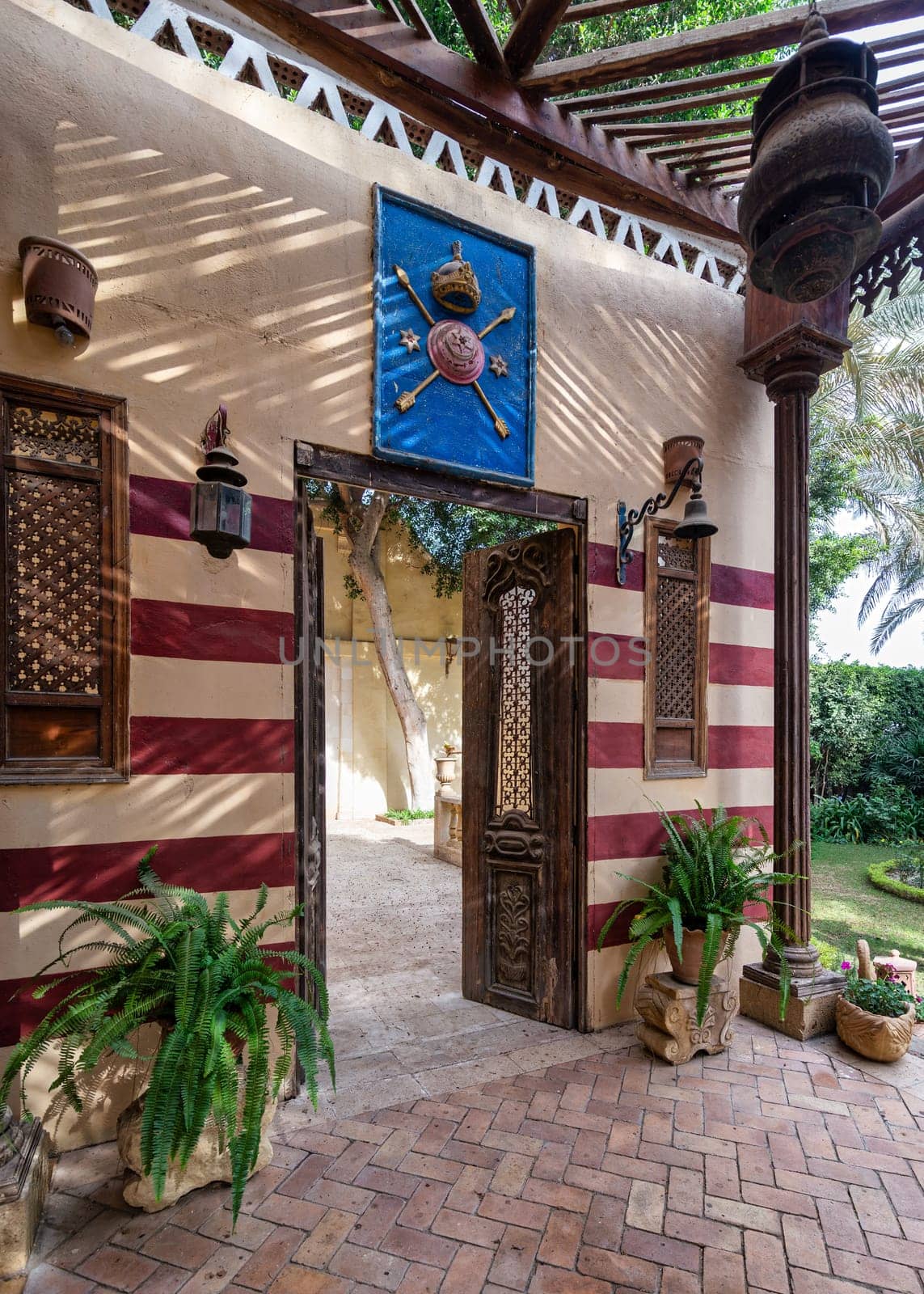 A beautifully crafted wooden door opens to a courtyard with red and white striped wall, wooden columns and lush greenery plant pots