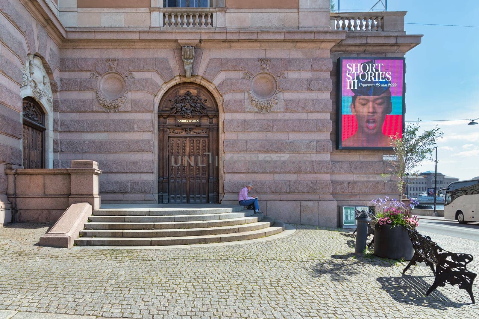 Stockholm, Sweden - June 26 2022: Elderly man reading a book at a staircase infront of brick stone wall with a billboard advertising Short Stories III and wooden door leading to the Royal Opera House