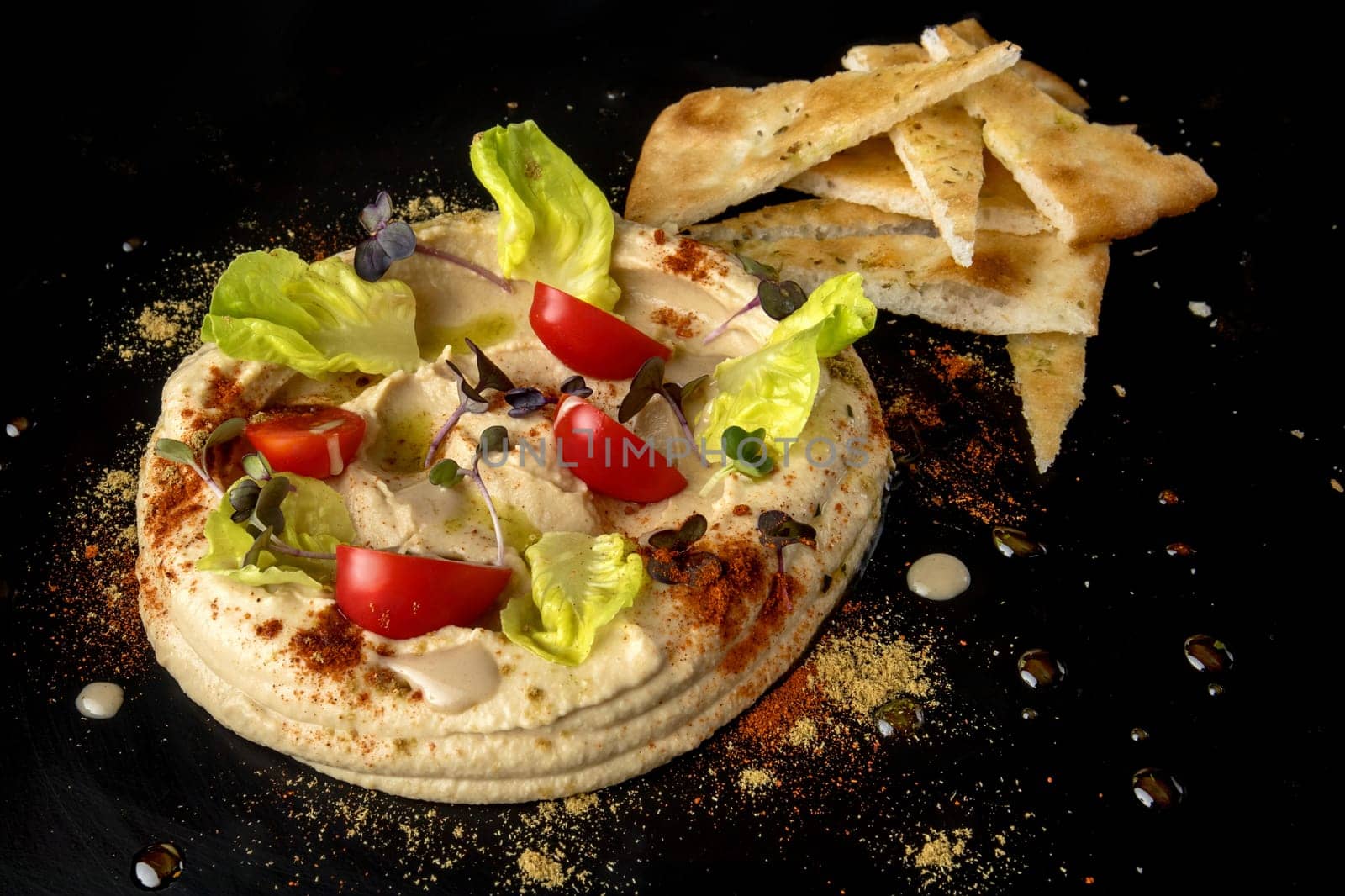 Indulge in a captivating hummus presentation, with smooth hummus forming a circular centerpiece adorned with juicy tomato slices, fresh green salad, and vibrant microgreens, all delicately dusted with paprika. Complemented by crispy pita bread slices, this platter promises a harmonious fusion of flavors and textures.