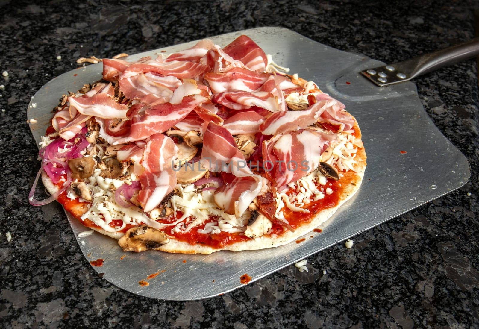 irresistible pizza topped with ham, mushrooms, cheese, onions, and ketchup, poised on a stainless steel peel on a granite table, ready for the oven. Get ready for a burst of flavors in every bite
