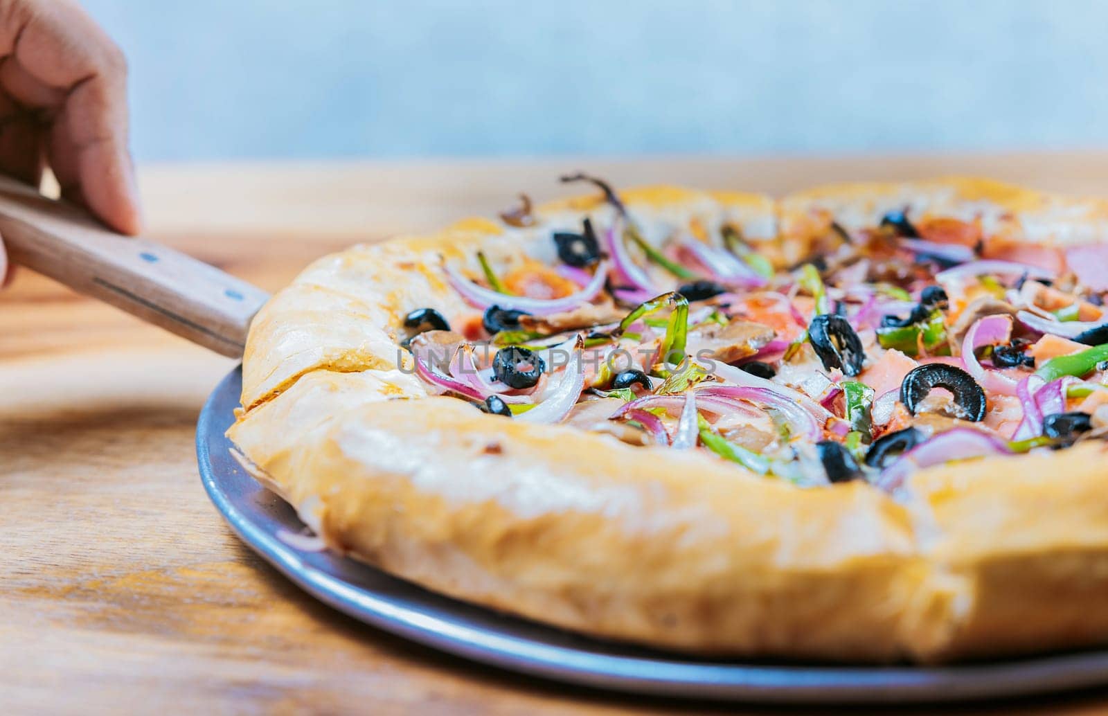 Hand taking a slice of supreme pizza with spatula. Close-up spatula taking a slice of supreme pizza with vegetables