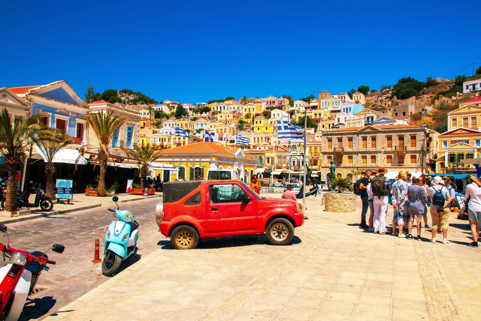SYMI, Greece - June 03, 2021. Tuorists at the harbour of Symi Town. Colorful villas stretch up the steep slopes.