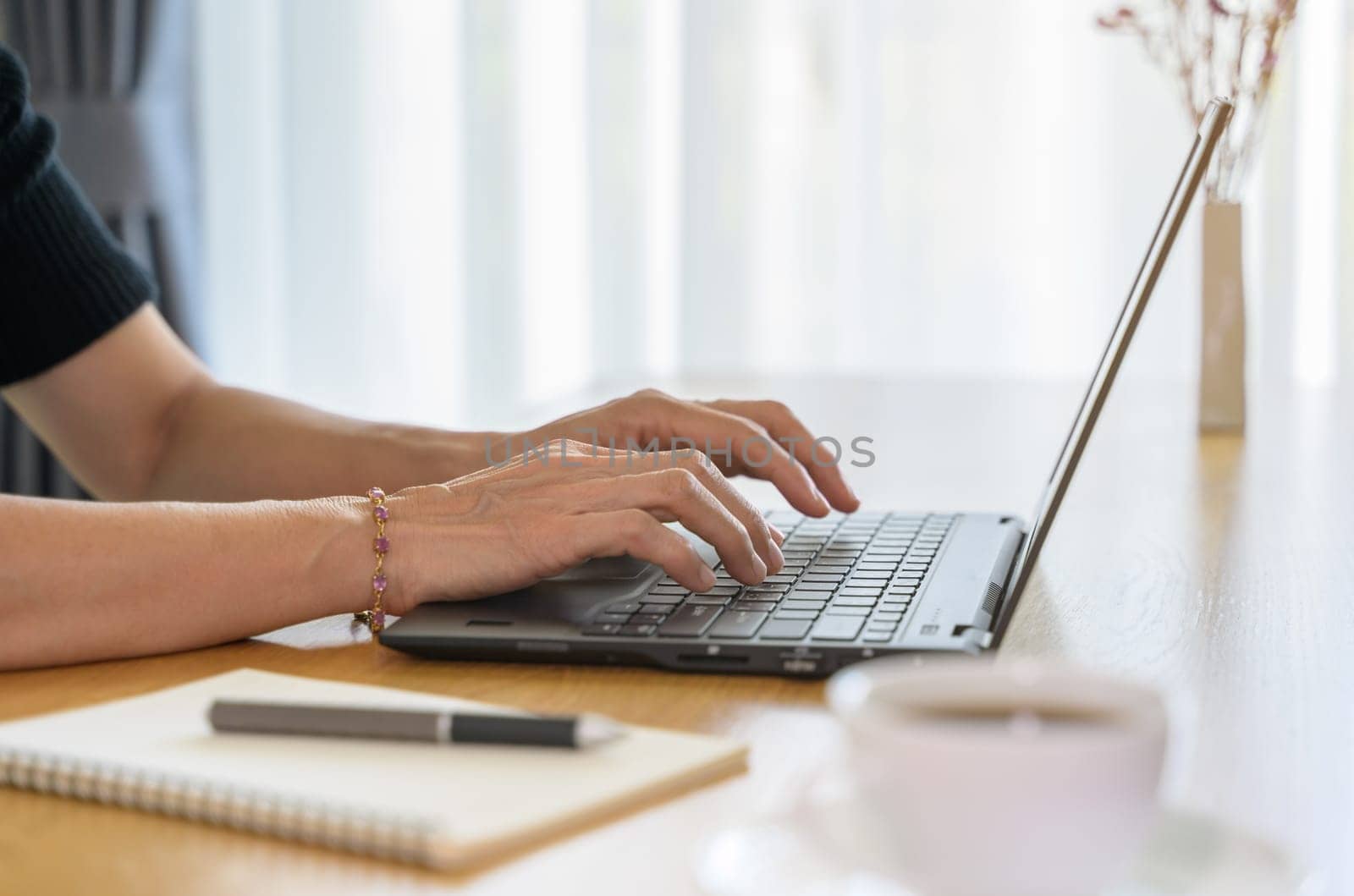 Close up on hands of female in causal cloth working on laptop computer at home while sitting at desk with coffee cup and notebook