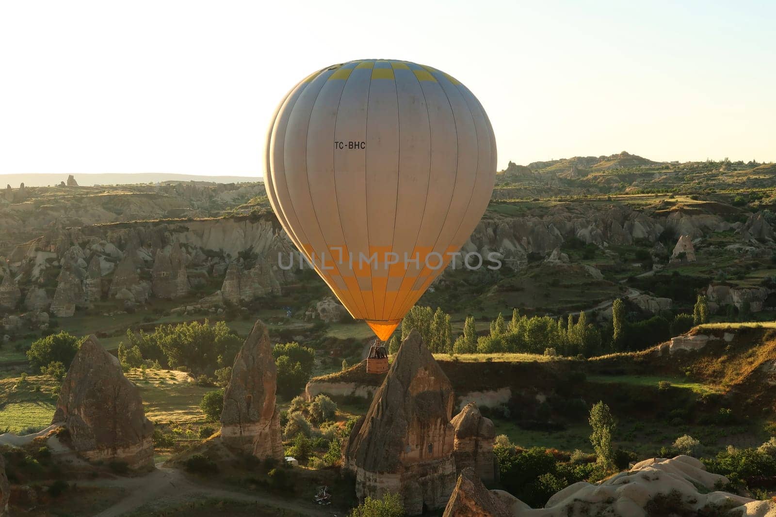Yellow and white hot air balloon illuminated by the early morning sun while flying over the landscape of Cappadocia with its famous fairy chimneys, close to Göreme, Cappadocia, Turkey 2022