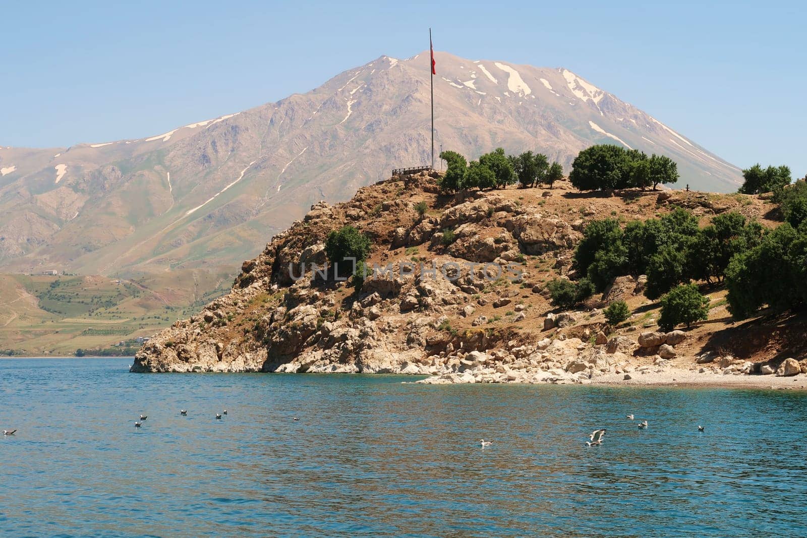 View from a boat onto Akdamar Island in the foreground with the red turkish flag on a pole and the snow covered Mount Artos in the background, Lake Van, Van Gölü, Turkey 2022