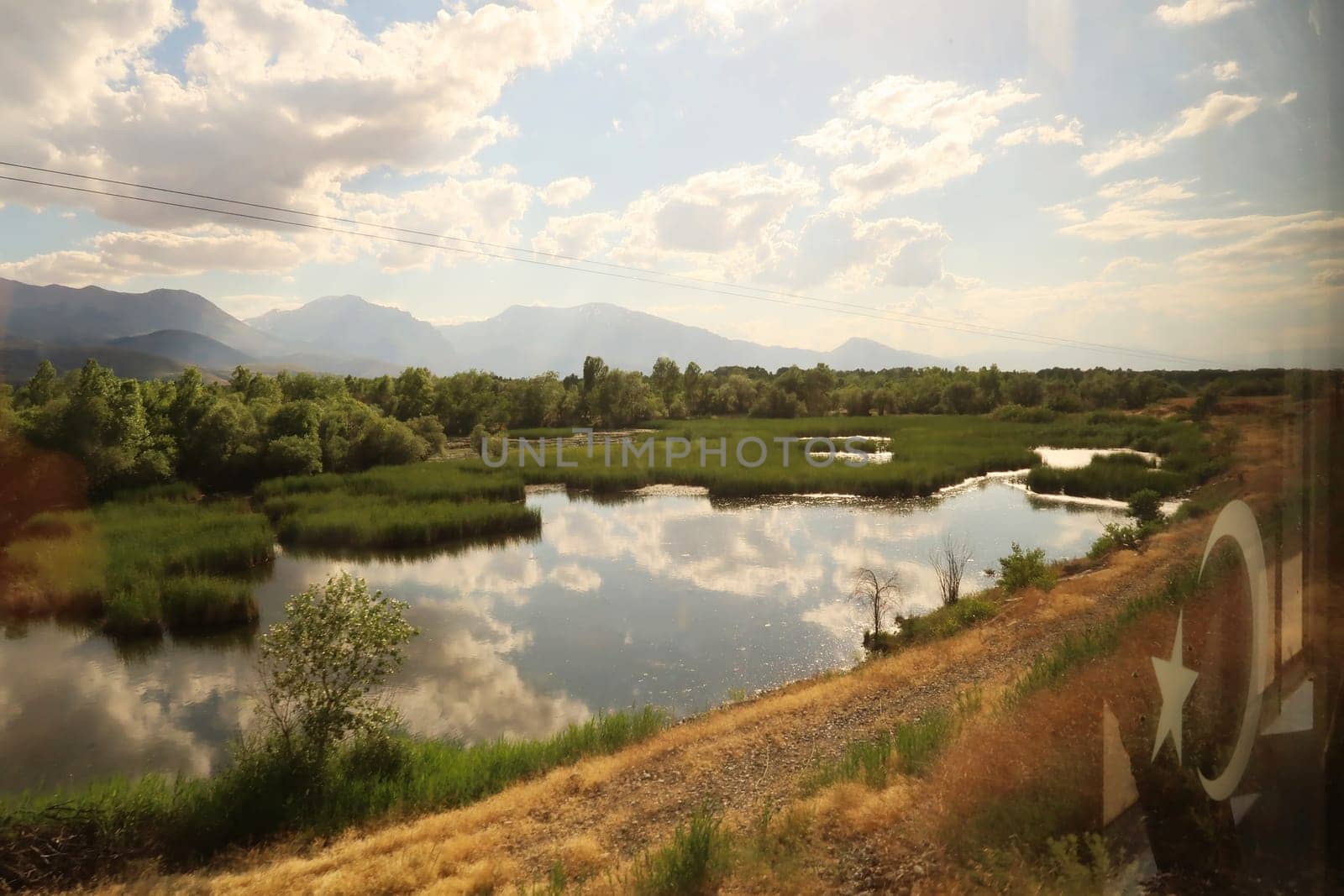 Beautiful landscape seen on the train ride in the Eastern Express, Dogu Ekspresi from Kars to Ankara, reflection of clouds in a pond, swamp surrounded by forest, mountains in the background, Turkey 2022