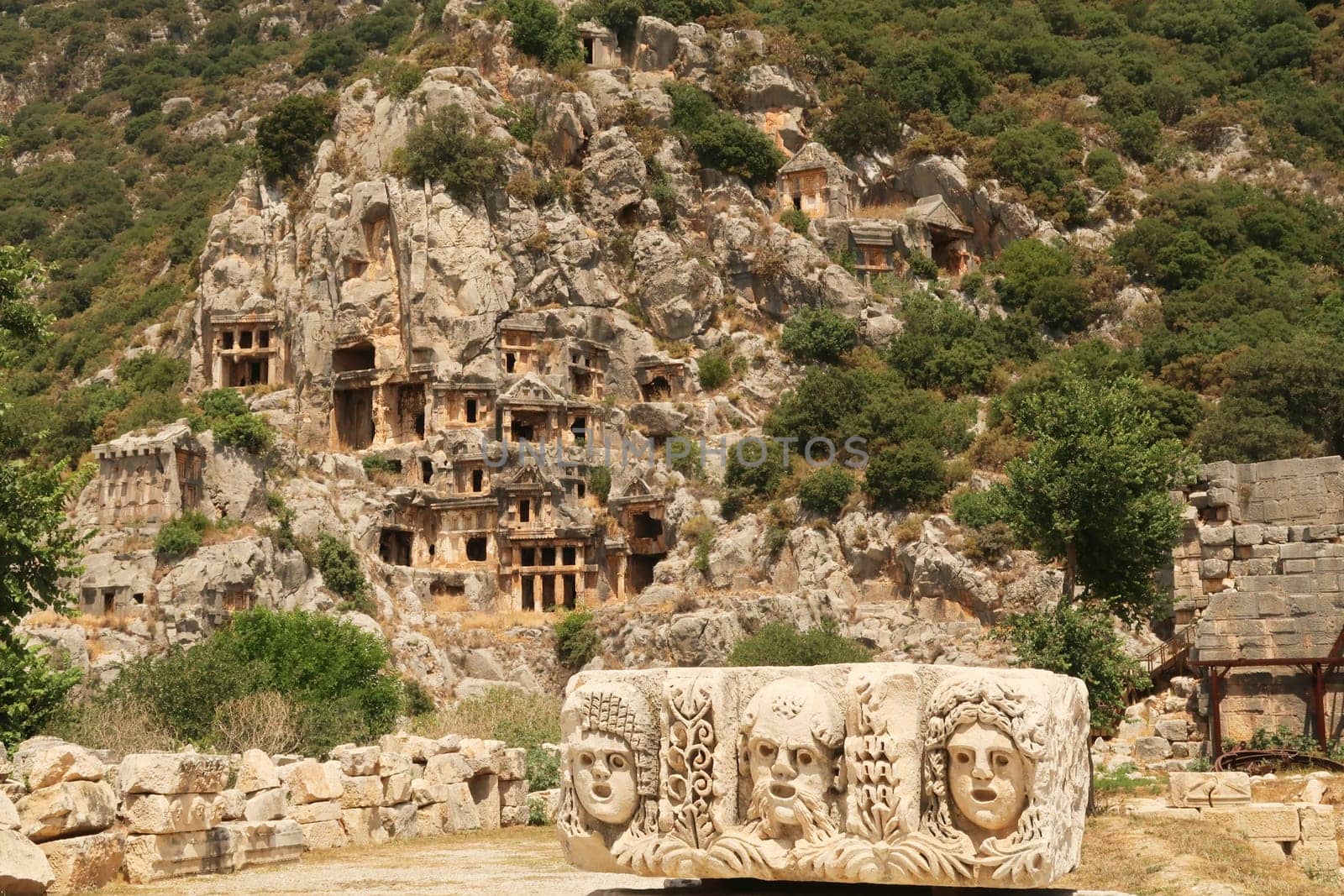 The ancient site of Myra with a stone relief with three theater masks in the front and the famous rock cut tombs in the background, near Demre, Turkey 2022