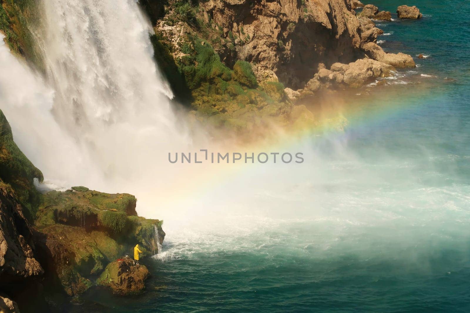 Fisherman in bright yellow raincoat trying to catch fish from a rock right next to the Düden Waterfalls in Antalya, Turkey 2022
