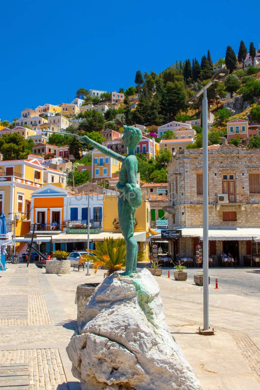 SYMI, Greece - JUN 03, 2021. Boy sculpture on Symi town promenade in the harbour in front of colorful villas that rise up the steep mountain slopes.