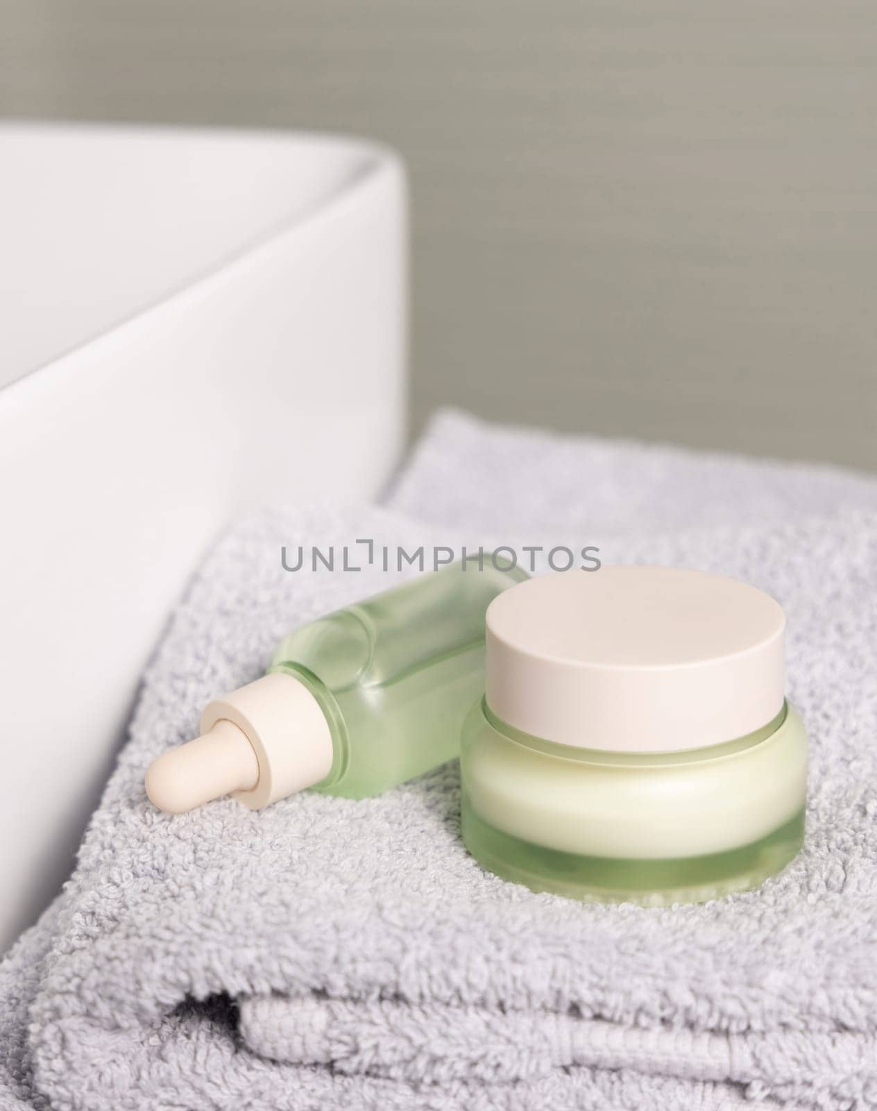 Translucent light green dropper bottle and closed cream jar on light grey folded bath towel near basin closeup, cosmetic mockup. Beauty and skincare routines with natural organic products