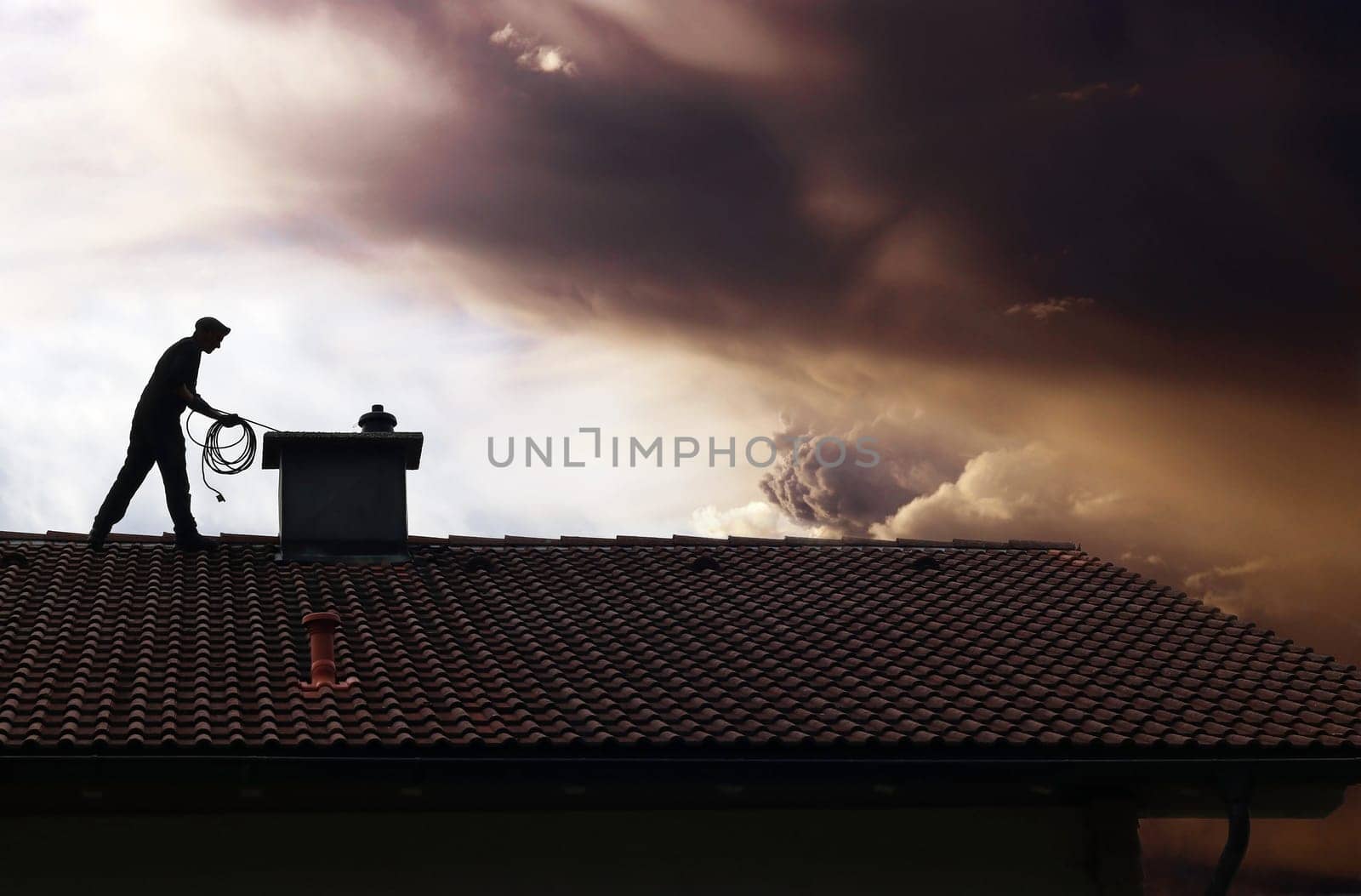 A chimney sweep cleans the chimney on a house roof while thunderclouds gather