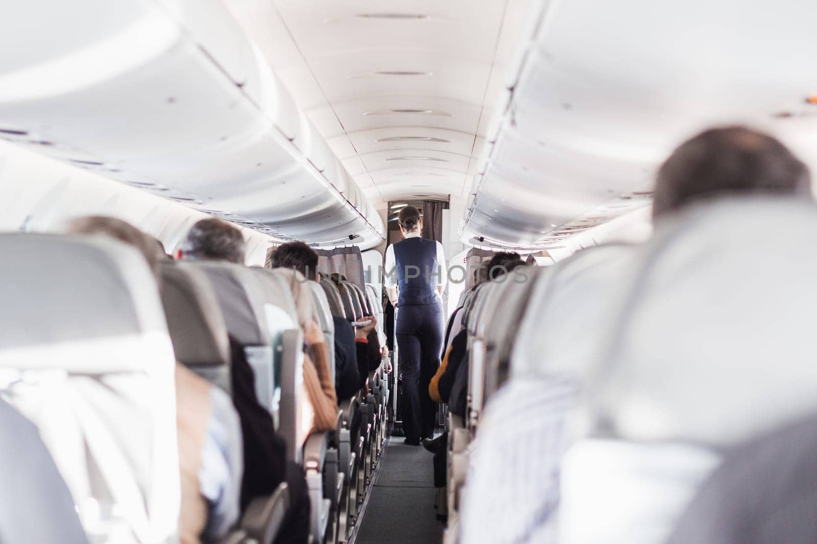 Interior of airplane with passengers on seats and stewardess in uniform walking the aisle, serving people. Commercial economy flight service concept