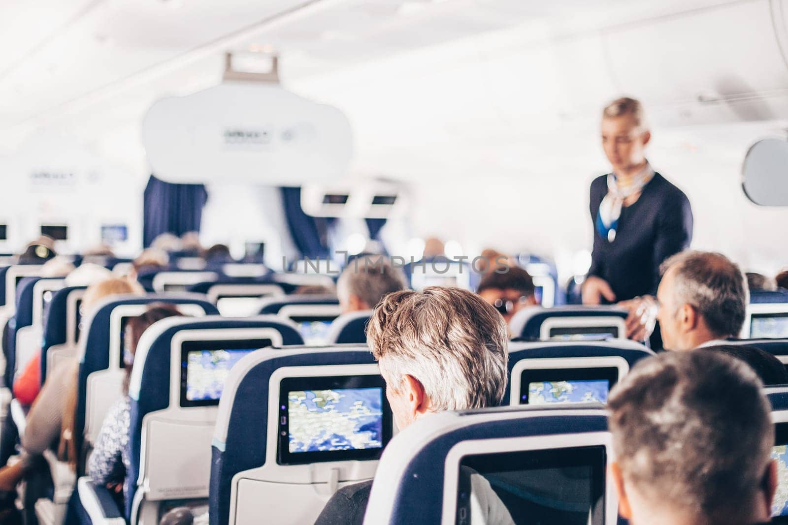 Interior of airplane with passengers on seats and stewardess in uniform walking the aisle, serving people. Commercial economy flight service concept
