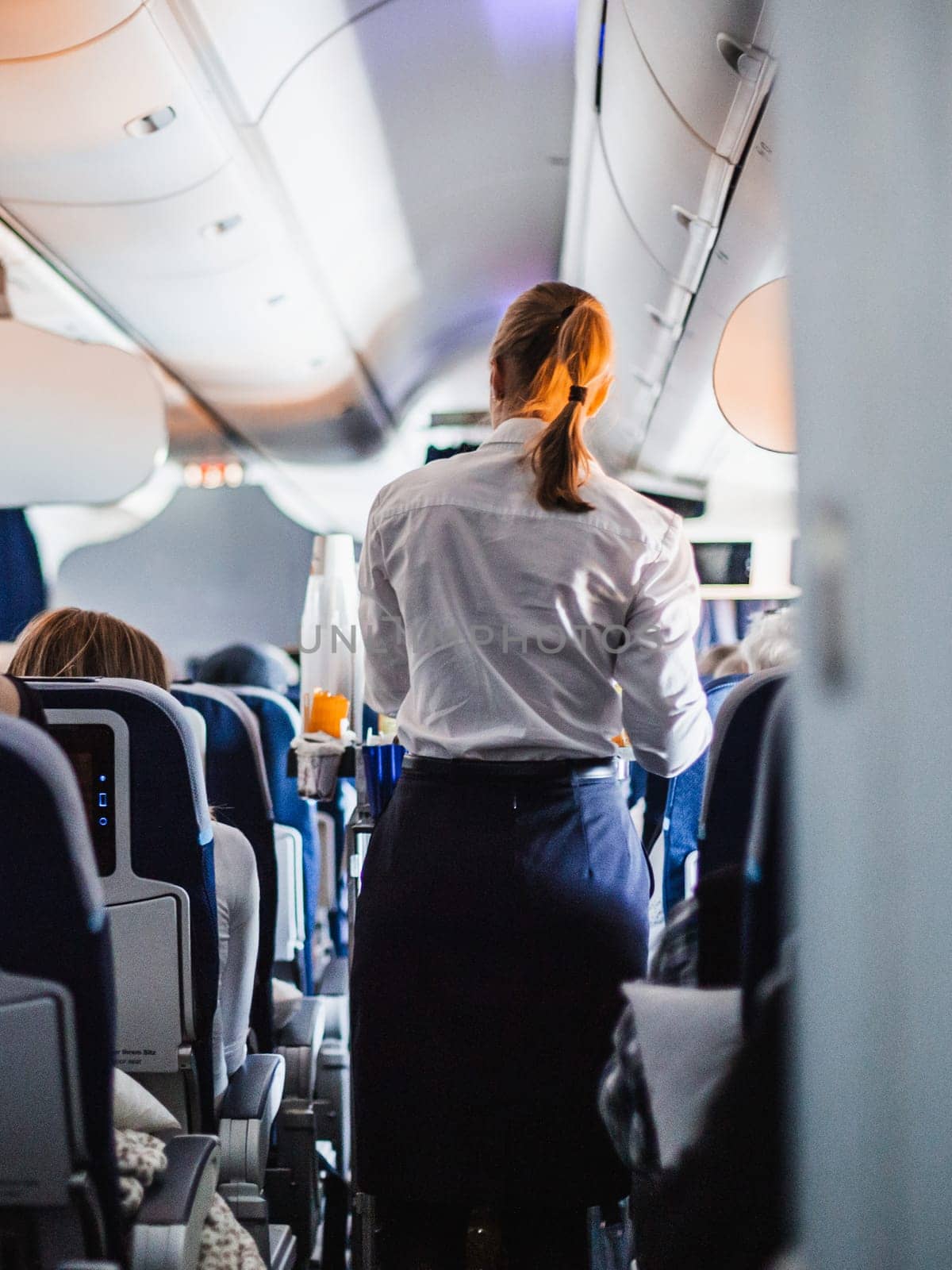 Interior of airplane with passengers on seats and stewardess in uniform walking the aisle, serving people. Commercial economy flight service concept