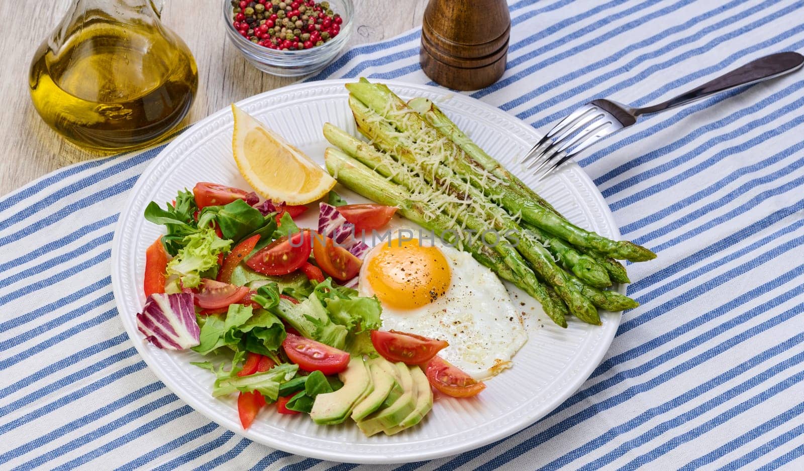 Round plate with cooked asparagus, fried egg, avocado and fresh vegetable salad on the table, top view