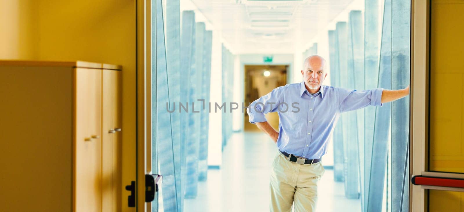 Standing in a bright hallway, an elderly man in a blue shirt leans against a door frame. He looks serious, thinking about future challenges like climate change, job losses, and power concentration