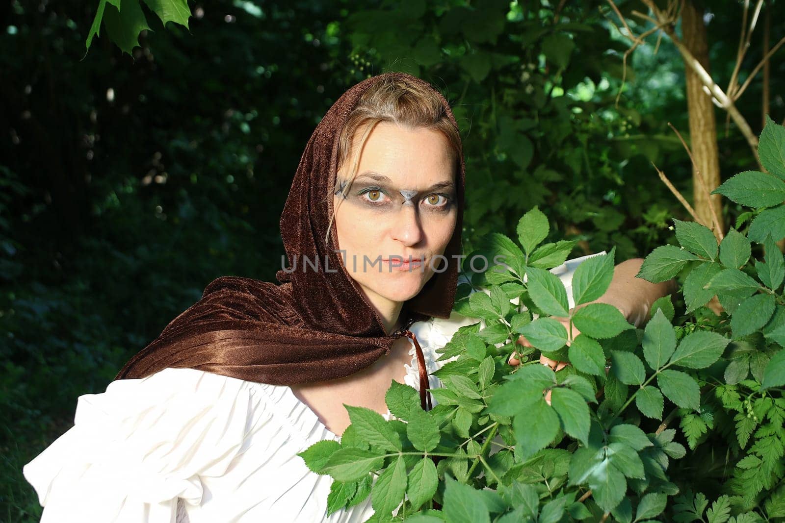 A woman as a medieval huntress in a dress with a hood hides in the forest