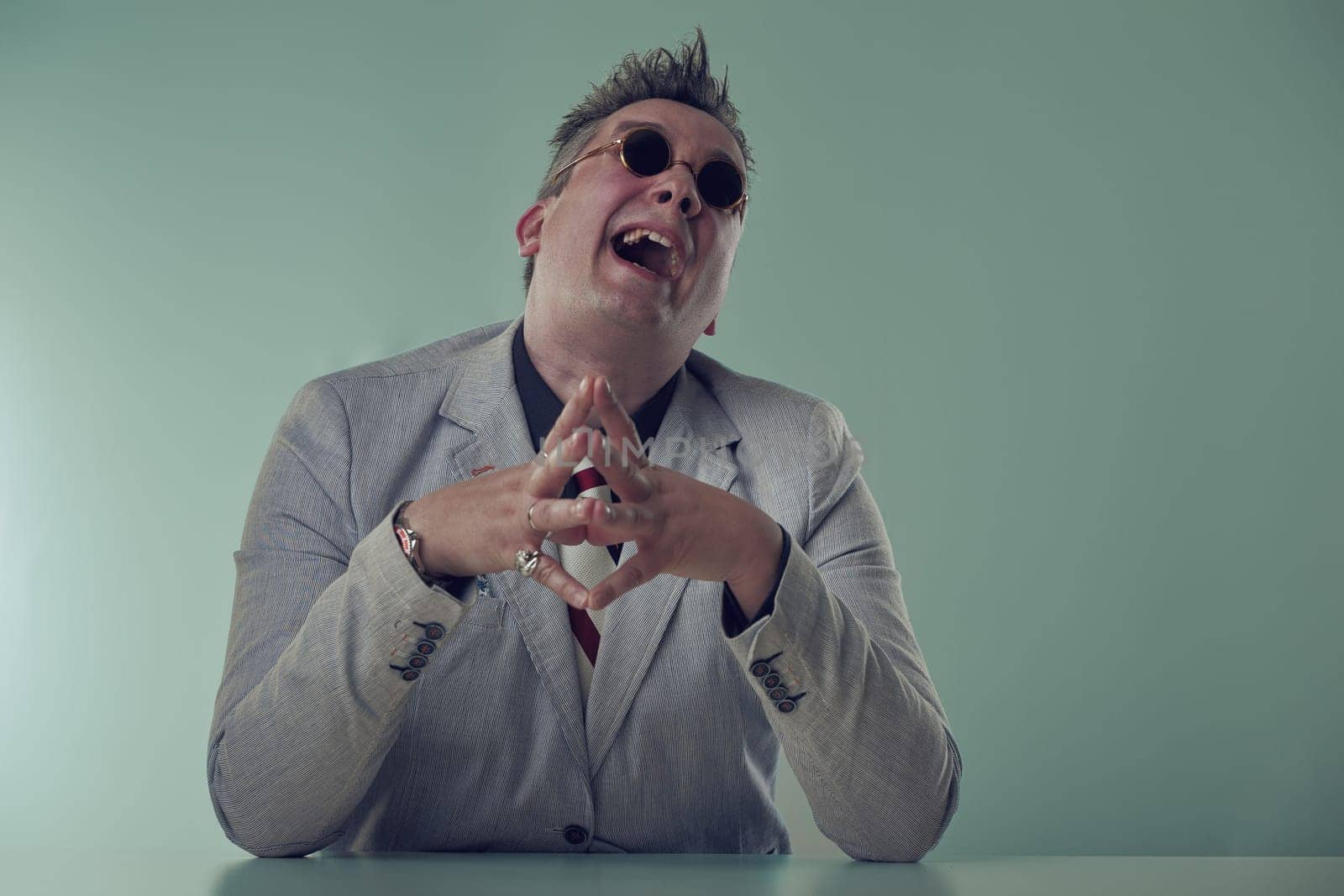Mad evil Joyful executive in grey blazer and sunglasses laughs uproariously, showing a spirited personality at his desk