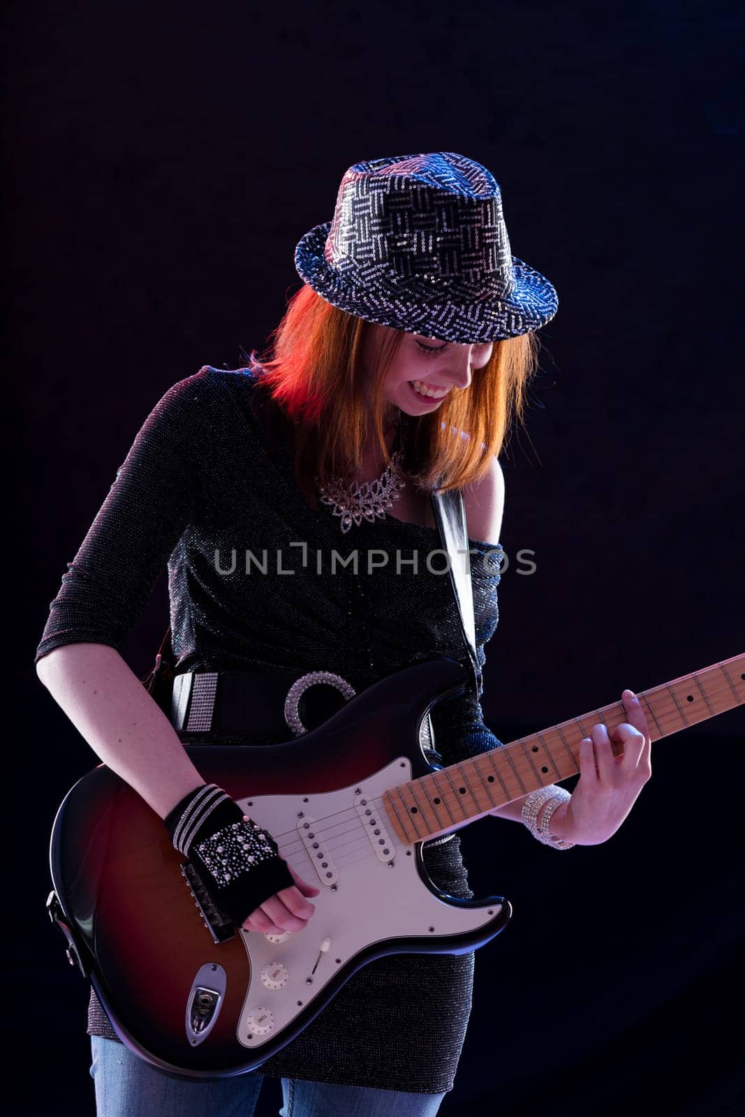 Smiling young woman with auburn hair plays an electric guitar on stage. She wears a sparkling hat, dark glittery top, jeans, and fingerless gloves, exuding confidence and joy in her performance