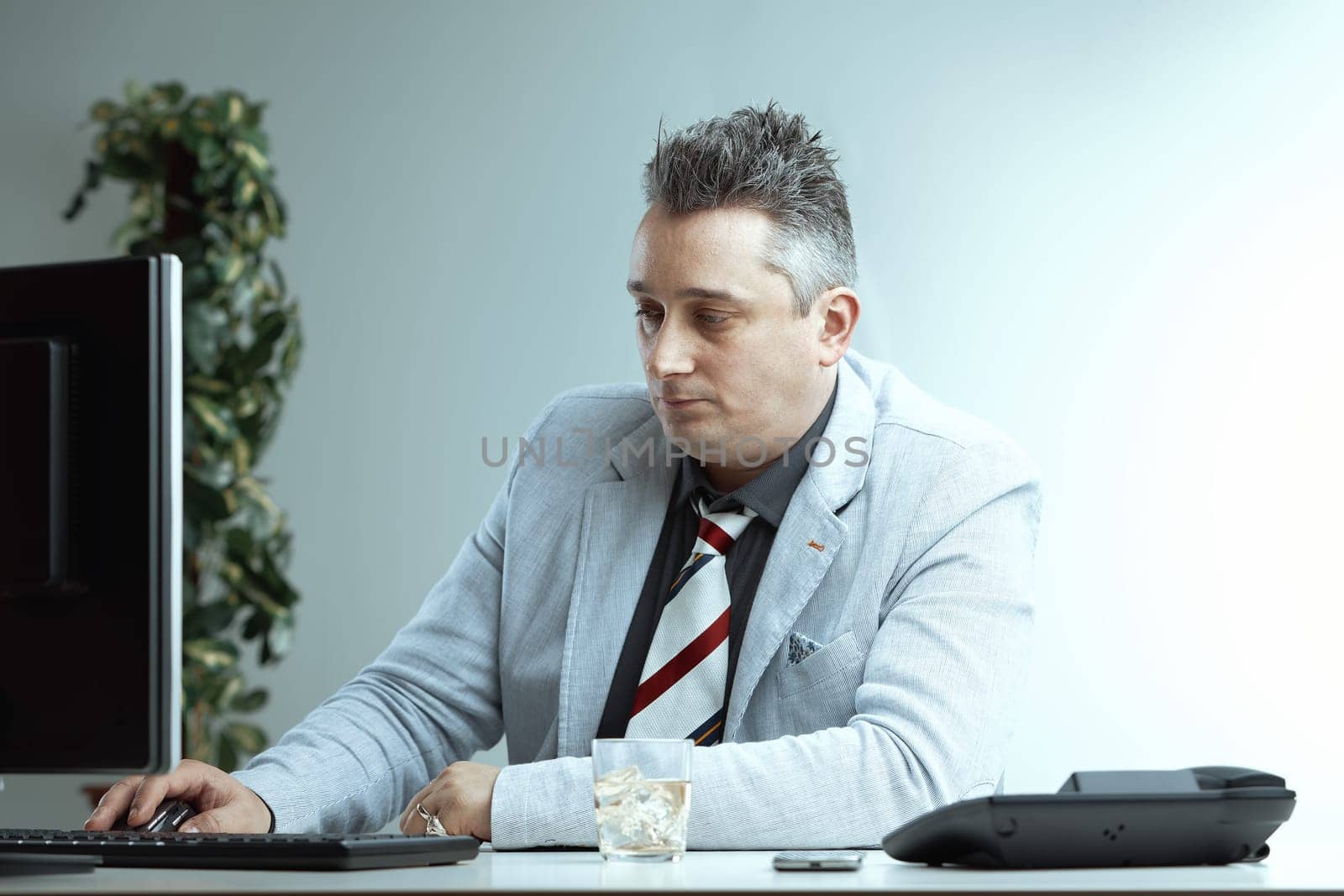 middle-aged man in a light suit and striped tie works at a computer with a glass of whiskey on the desk. His focused expression contrasts with the casual presence of alcohol at work