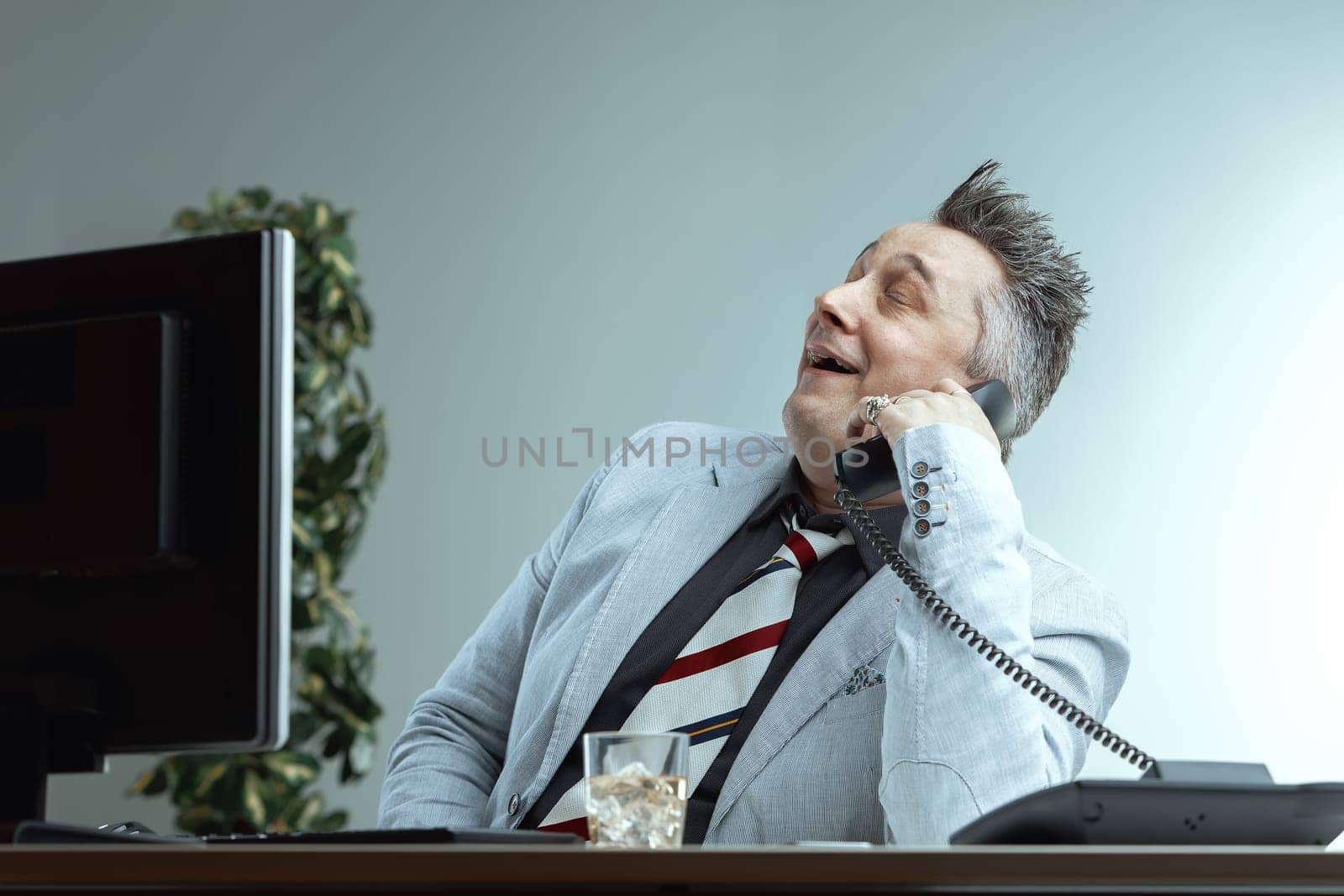 middle-aged man in a light suit and striped tie leans back, smiling confidently while talking on the phone. His smug expression and the whiskey glass on his desk suggest he is boasting about his knowl