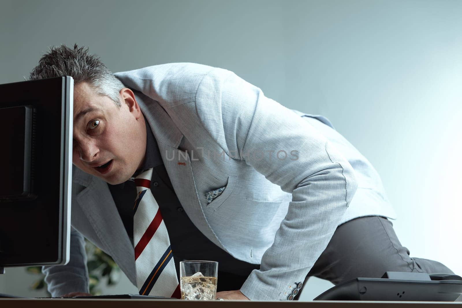man, dressed in a light suit and striped tie, appears startled while leaning over his desk. His surprised expression and awkward posture, along with a whiskey glass, imply he was caught doing somethin
