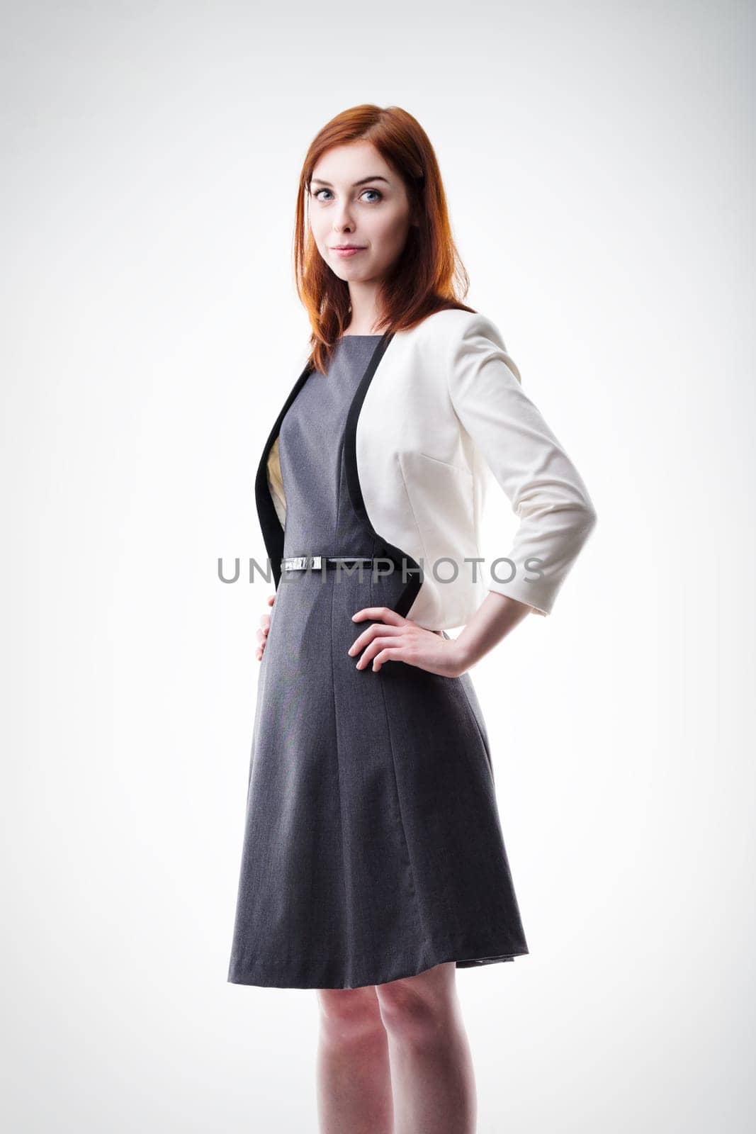 young woman with auburn hair, dressed in a white blazer and dark dress, stands confidently with hands on hips. Her poised demeanor and professional attire convey authority and competence