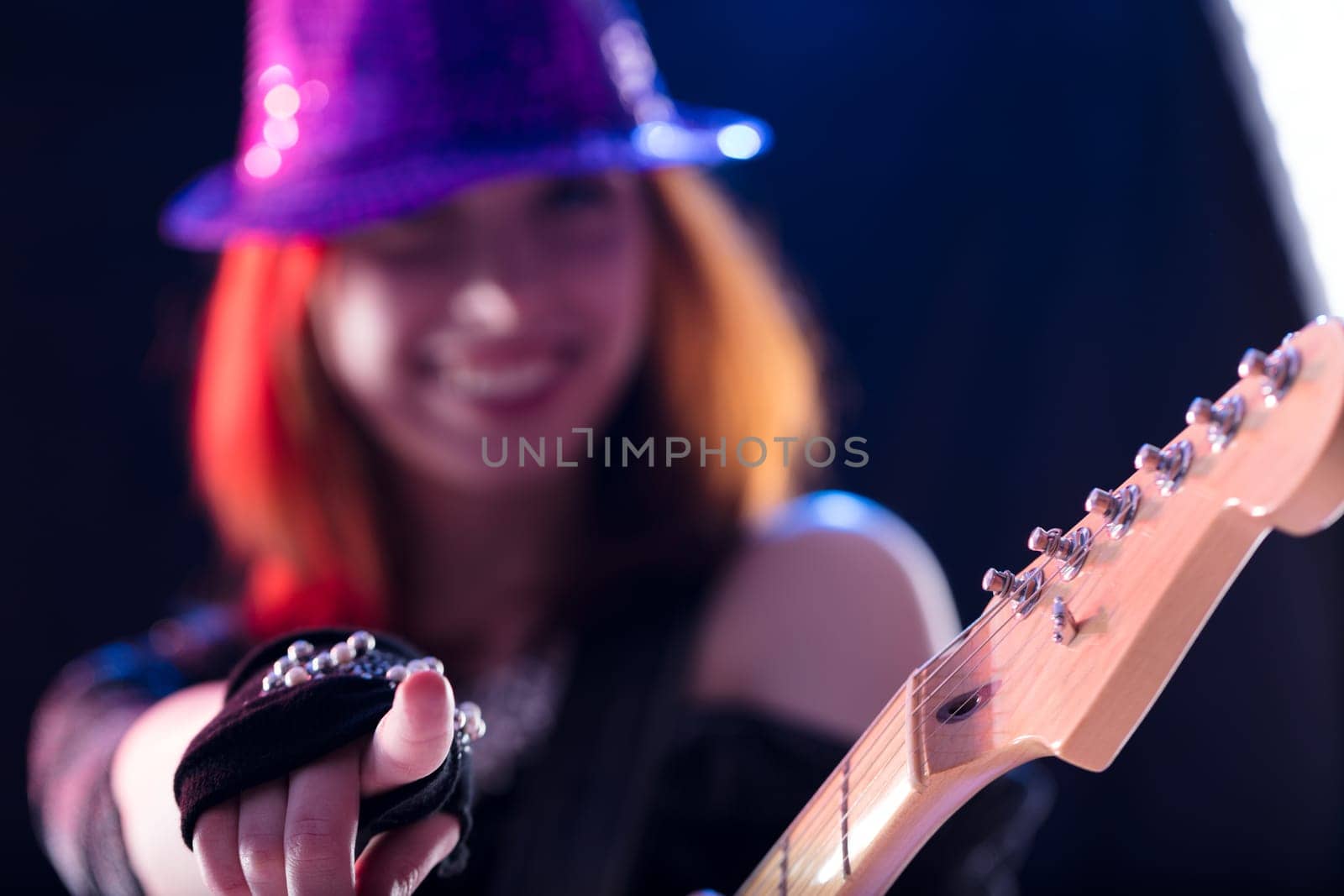 Young woman with auburn hair, sparkling hat, and dark glittery top plays an electric guitar with a smile. Her confident and joyful performance on stage showcases her musical talent