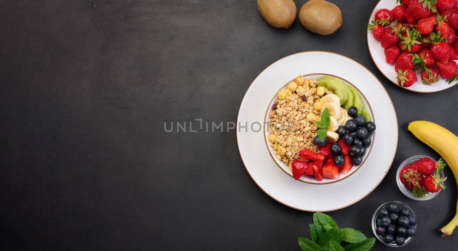 Granola with strawberries, kiwi, banana and blueberries in a round plate on a black table. Healthy and tasty food, top view. Copy space