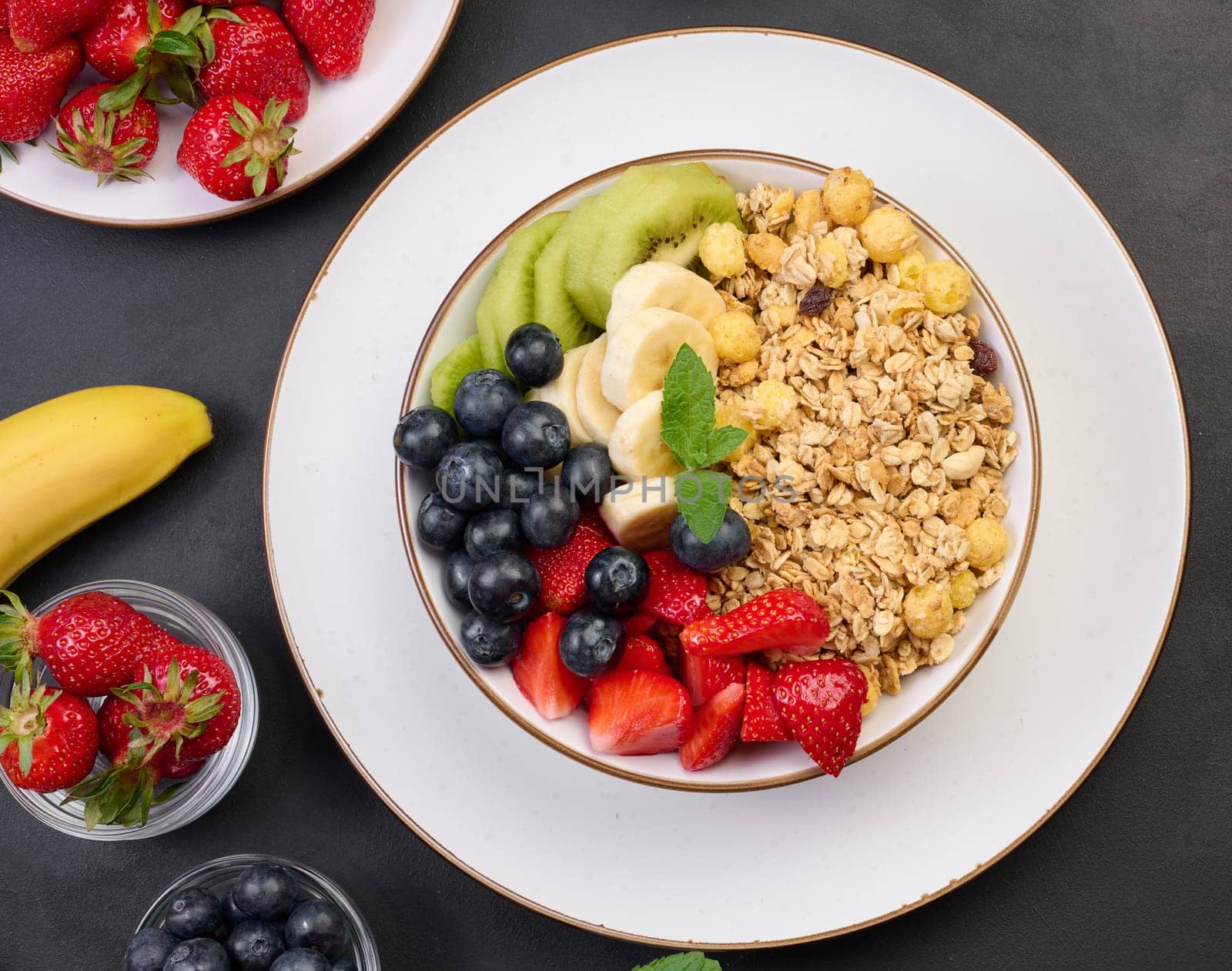Granola with strawberries, kiwi, banana and blueberries in a round plate on a black table. Healthy and tasty food, top view