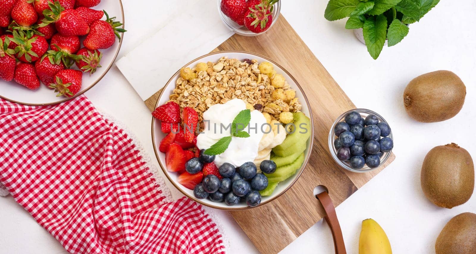 Granola with strawberries, kiwi, banana and blueberries in a round plate on a white table. Healthy and tasty food, top view