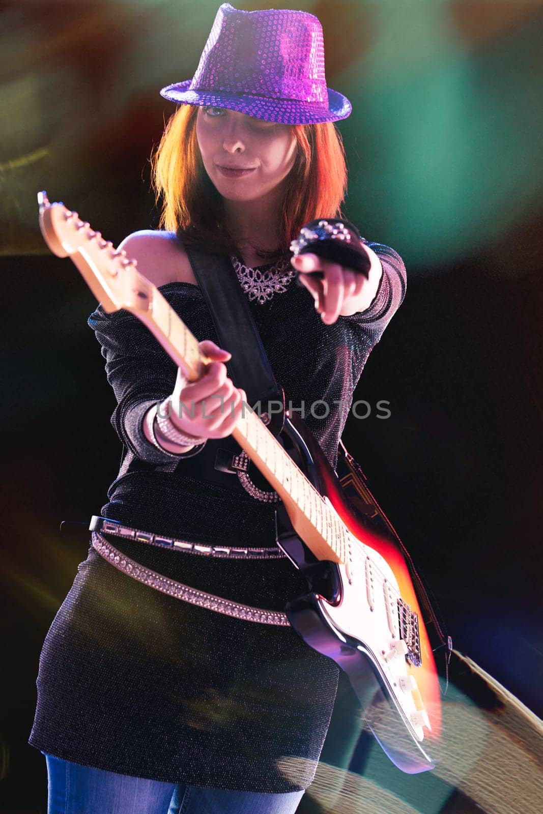 joyful young woman with red hair, in a glittering black ensemble and sequined hat, skillfully plays her electric guitar, stage lights casting colorful reflections around her