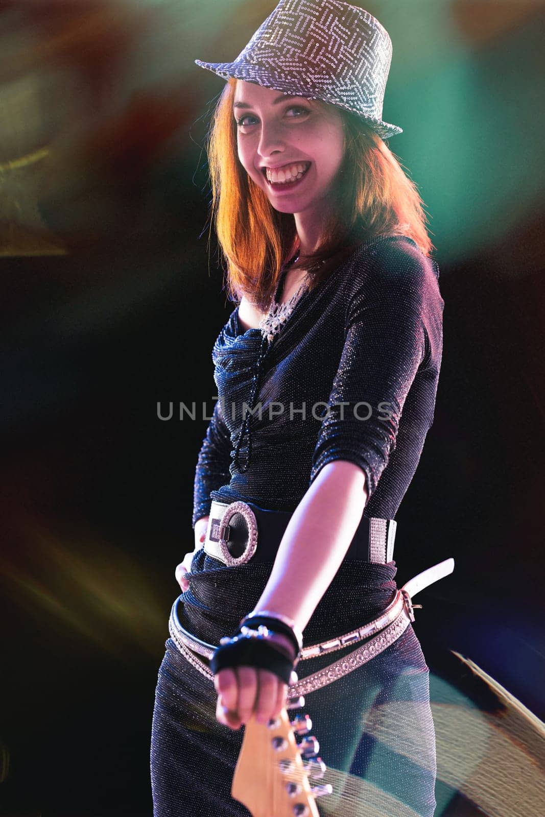 With a radiant smile, a red-haired young woman in a sparkling black outfit and sequined hat plays an electric guitar, her joy evident as colorful stage lights illuminate her