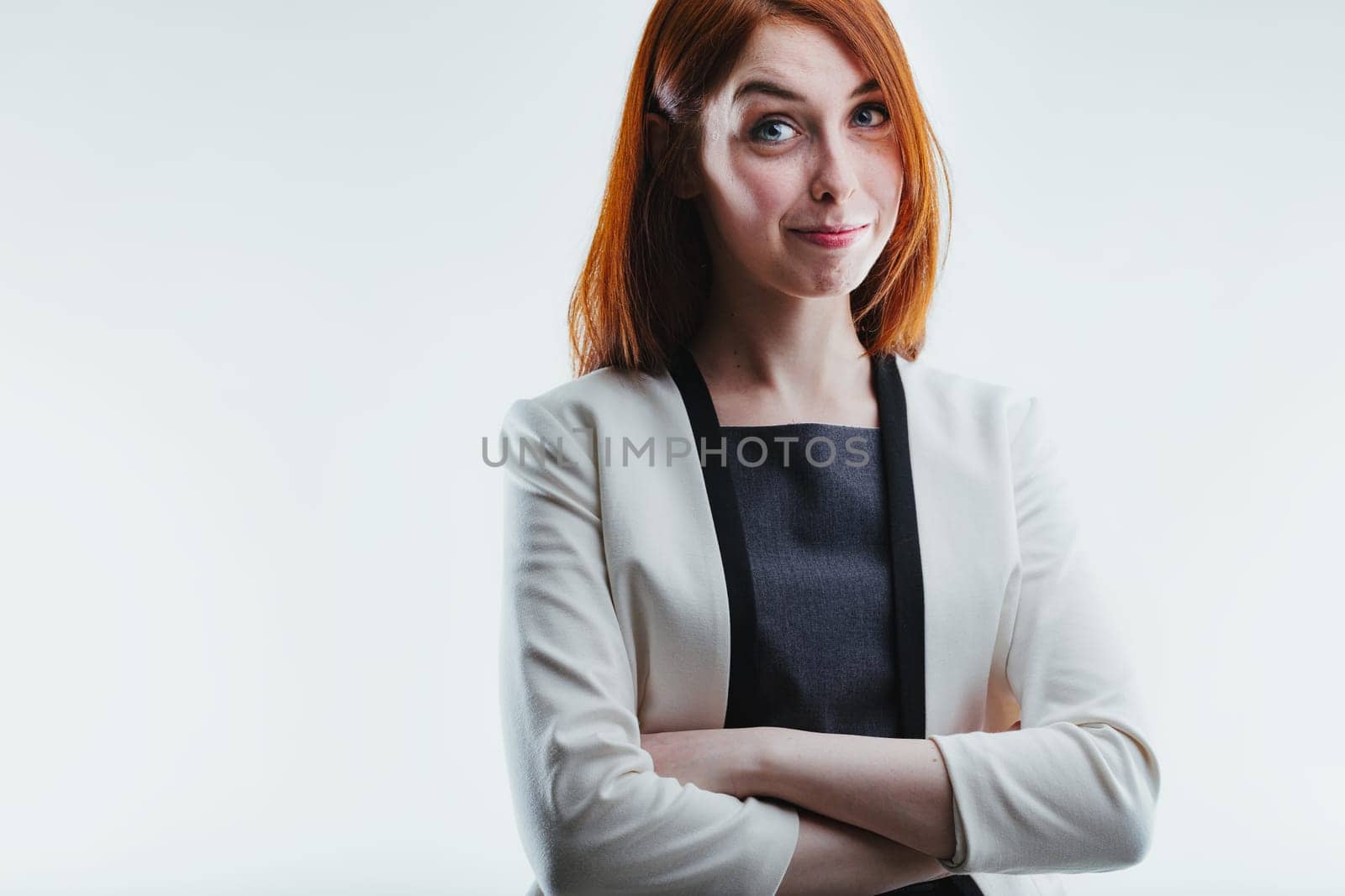 Young redhead displaying a subtle smirk, confidently standing with her arms crossed in a light gray suit jacket