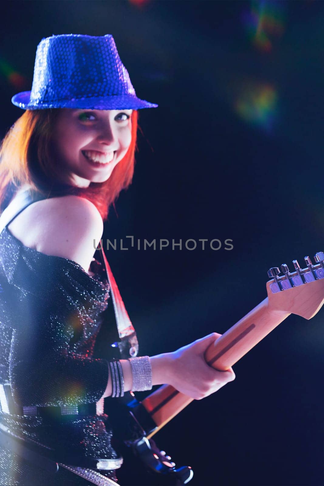 Enthusiastically playing her electric guitar, a young woman with red hair and a sequined hat glows under the stage lights, her black outfit shimmering with every movement