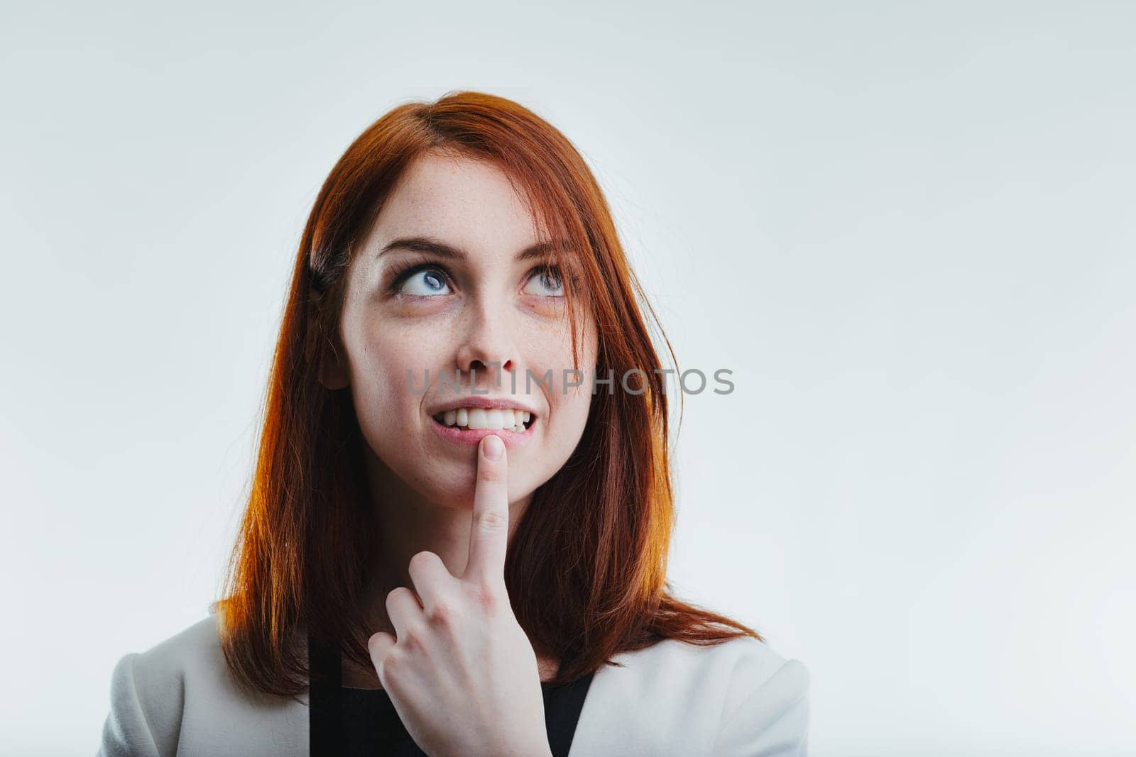 Thoughtful redhead with an inquisitive expression, gently touching her chin, dressed in a sophisticated white blazer