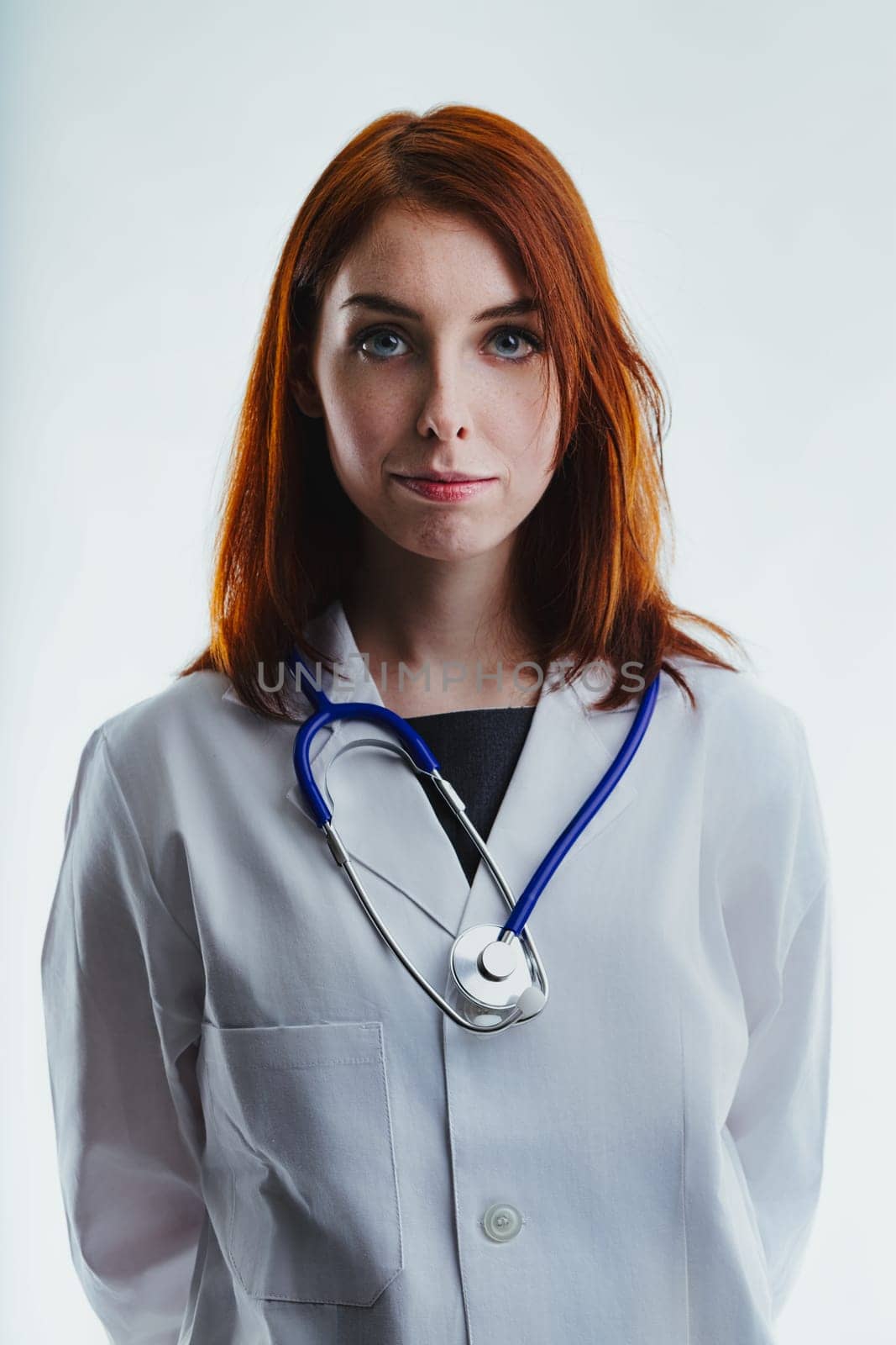 red-haired female doctor wearing a white lab coat and a stethoscope around her neck looks directly at the camera. Her serious expression highlights her professionalism and dedication to patient care