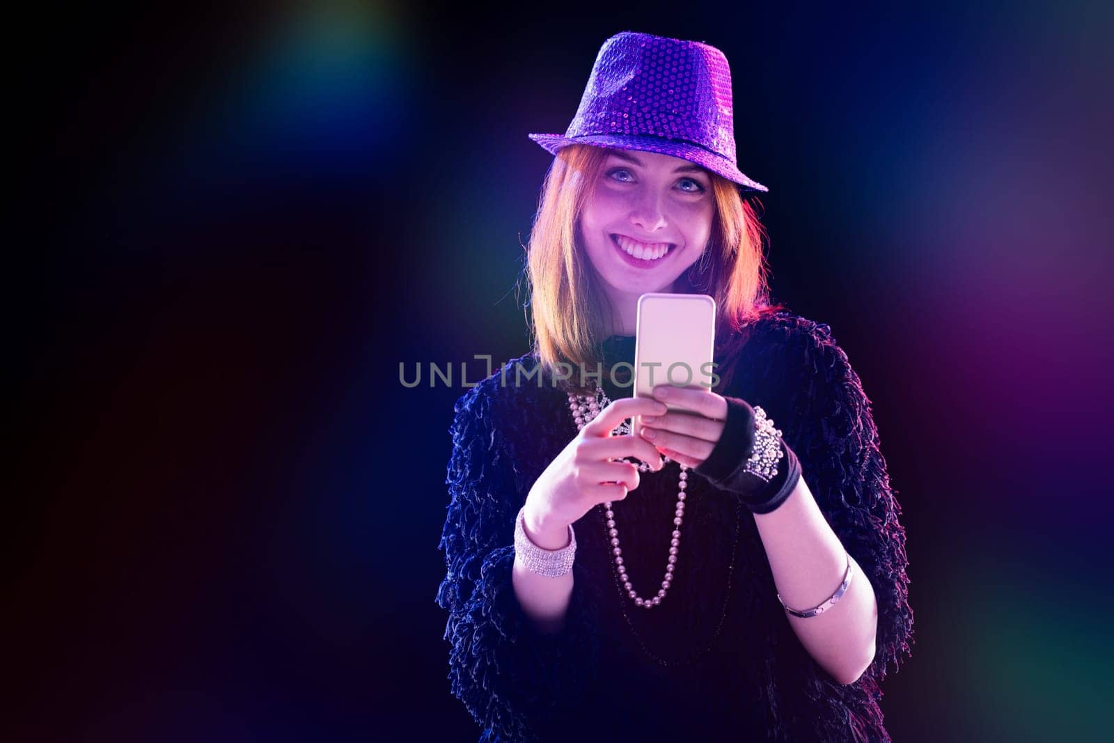 red-haired young woman in a purple sequined hat and sunglasses uses her phone, smiling confidently. Wearing a stylish black top and jeans, she is surrounded by stage lights and fog