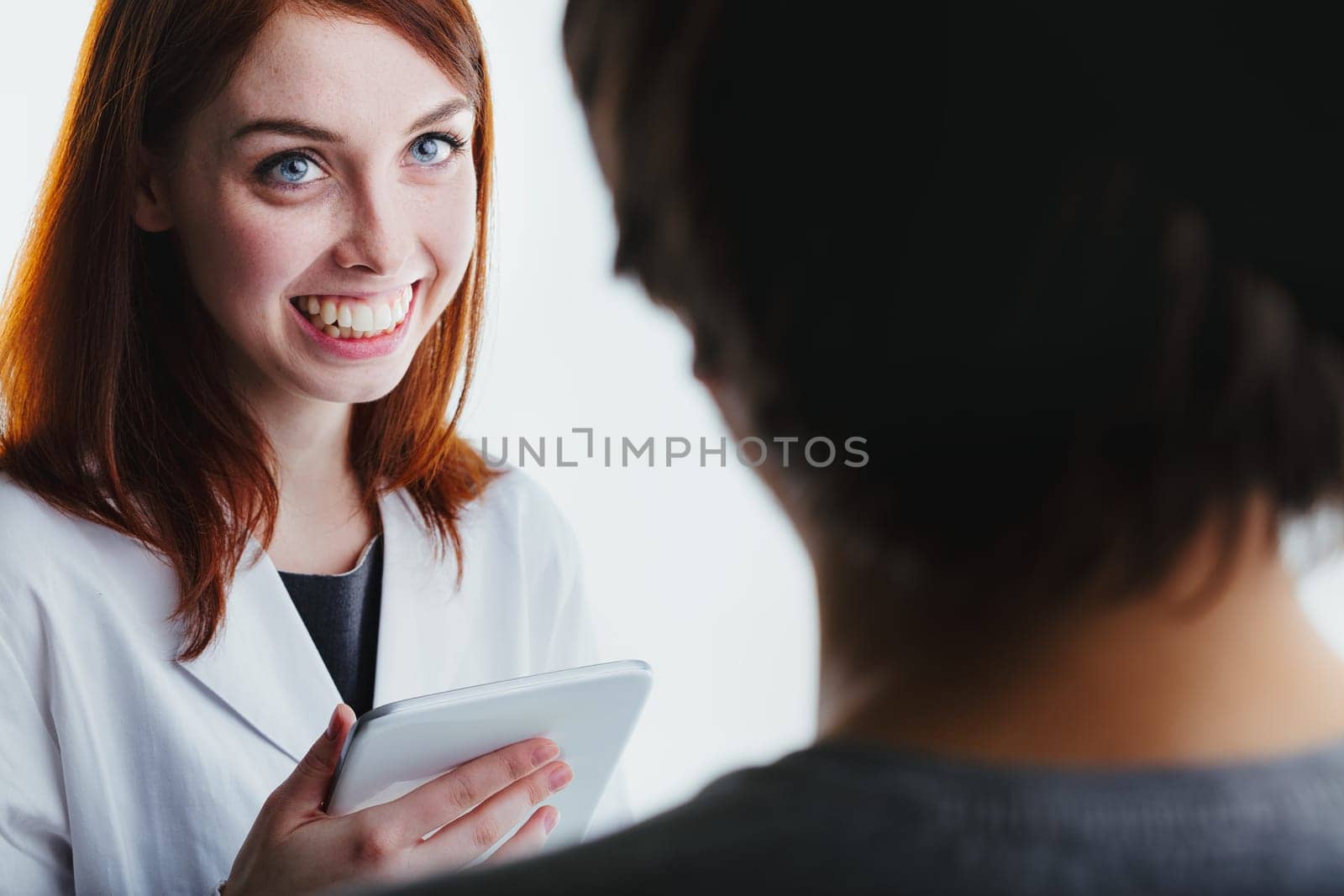 Smiling brightly, a red-haired woman in a lab coat uses a tablet while interacting with a patient, emphasizing the role of technology and empathy in modern medical practice