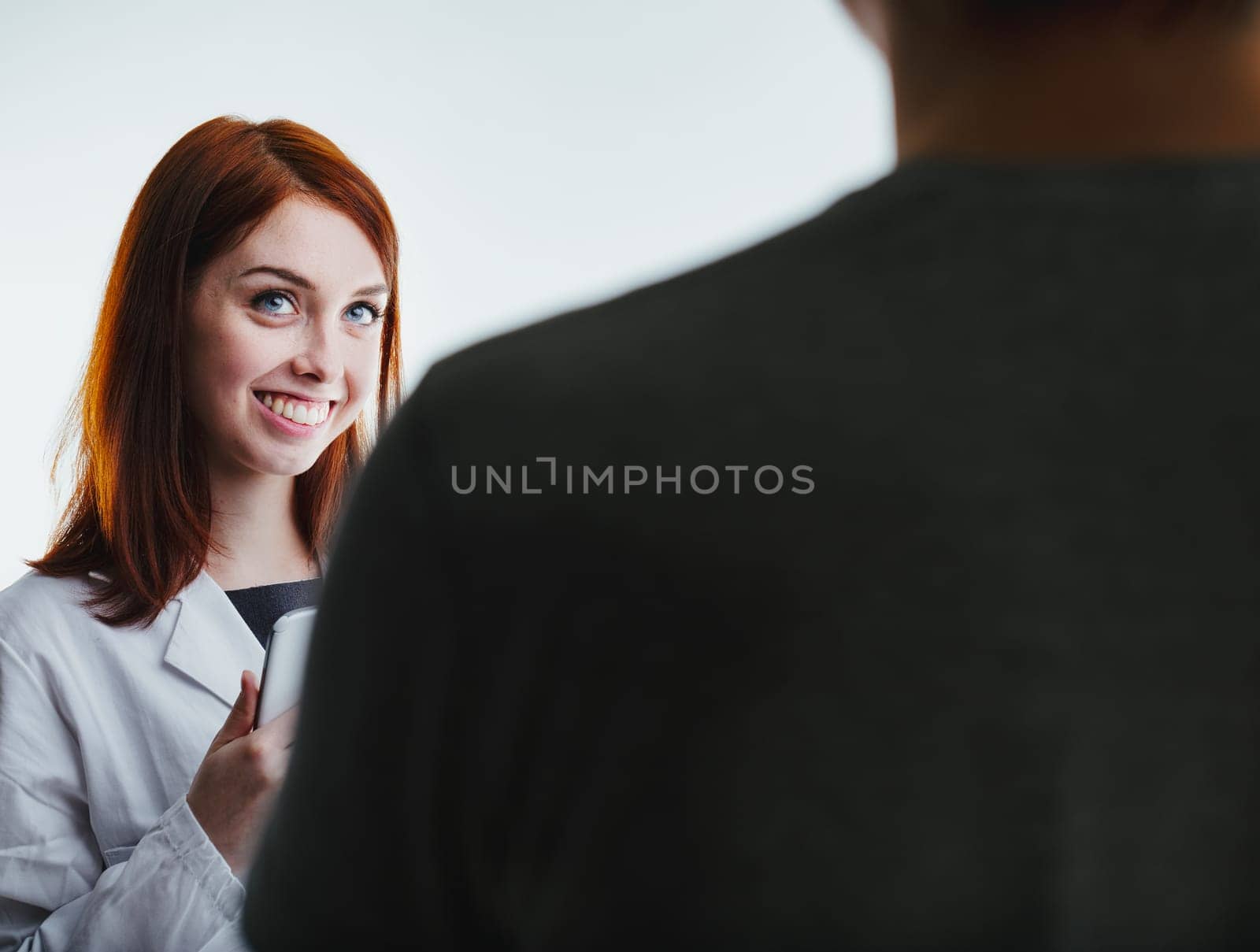 ed-haired female doctor in a white lab coat smiles warmly while holding a tablet, engaging with a patient. Her professional yet friendly demeanor underscores the importance of compassionate healthcare